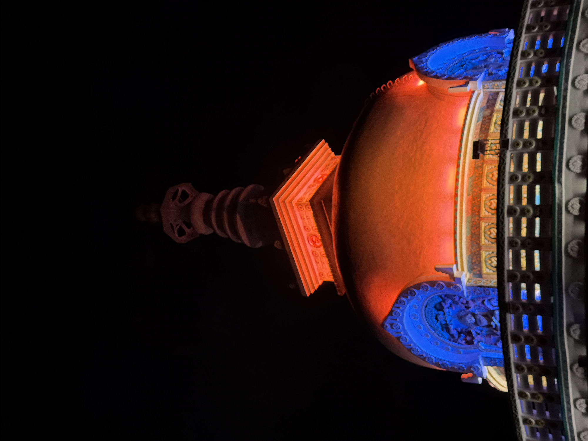 The image shows the Shanti Stupa in Leh, India, illuminated at night. The stupa is a Buddhist monument with a dome-shaped structure, topped with intricate carvings and golden spires. The structure is lit with vibrant blue and orange lights, highlighting its architectural details. The background is a dark night sky, emphasizing the illuminated stupa.