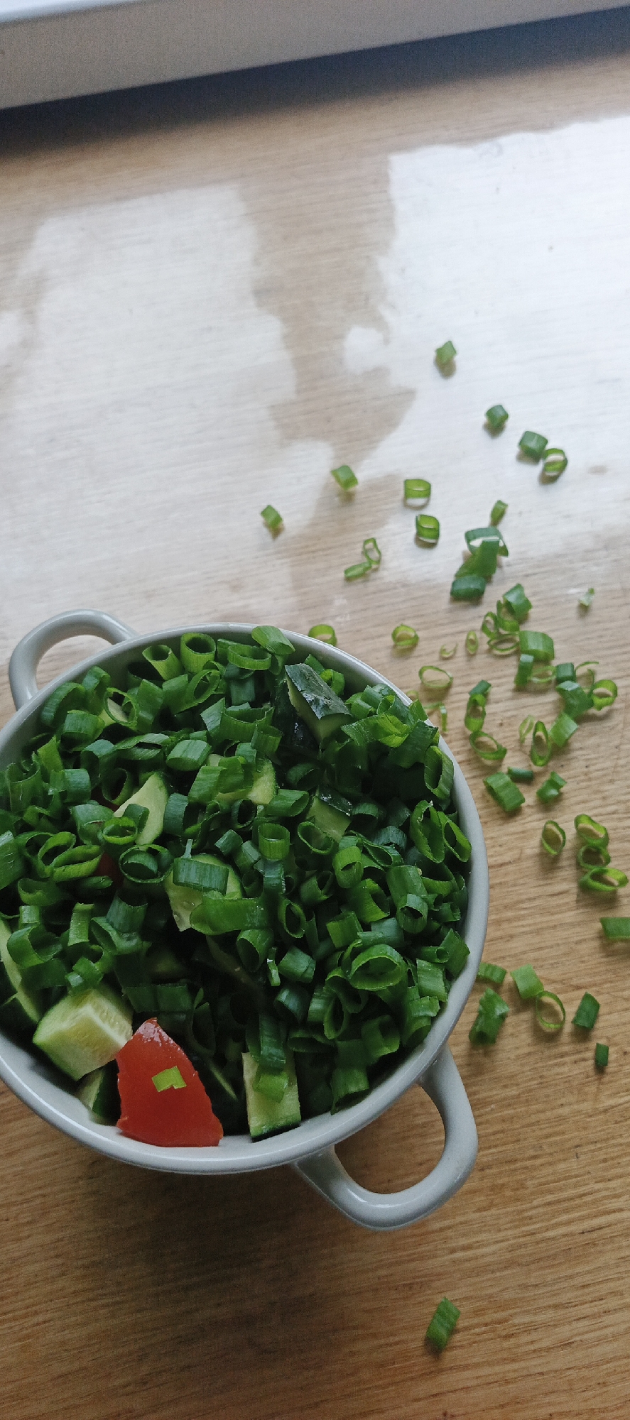 A light-colored, two-handled ceramic bowl, filled with chopped green onions and diced cucumbers, is centrally positioned on a wooden surface. A single triangular piece of red tomato is visible amongst the green ingredients within the bowl. Numerous fragments of chopped green onion are scattered across the wooden surface to the right and above the bowl. The wooden surface displays a subtle grain pattern. A dark, horizontal line is present at the top edge of the frame. No individuals are present. This scene is located in the city of Chaikovsky, Russia.