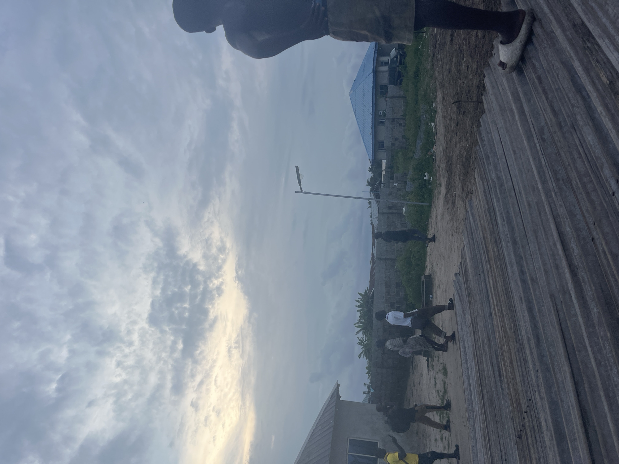 The image depicts an outdoor scene with several individuals. The perspective is from a low angle, and the image is rotated, with the horizon appearing on the left side.
In the foreground, a person is visible from the waist down, wearing shorts and a white slipper on their right foot. They are standing on a corrugated metal surface.
Further back, a group of people are scattered across a dusty, unpaved area. Some appear to be walking or standing. One person in the middle ground is wearing a white shirt.
In the background, there are several unfinished buildings. One building has a prominent blue-pitched roof. A street lamp is visible on a pole. Vegetation, including what appear to be banana trees, is present. A dark-colored vehicle is parked near one of the buildings.
The sky is overcast with scattered clouds, suggesting it might be daytime, possibly late afternoon or early morning due to the diffused light. The overall impression is that of a developing area or a construction site. No text is visible in the image.