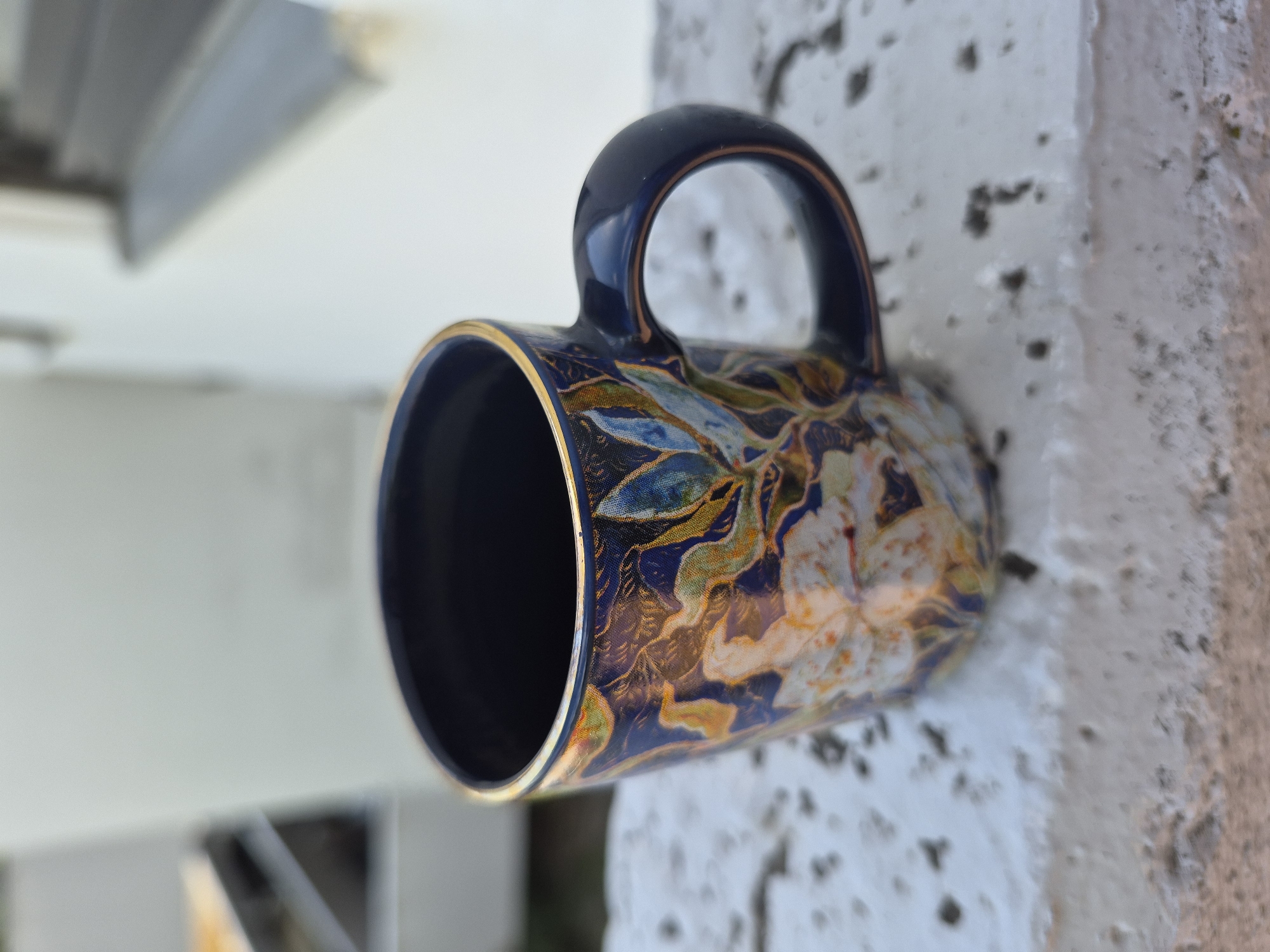 A close-up, sideways shot shows a dark blue ceramic mug with a floral design and a gold rim. The mug has a dark blue handle and is decorated with intricate patterns of flowers and leaves in shades of blue, gold, and white. The mug appears to be resting on or against a textured, light-colored surface, possibly a wall or pillar, which has a rough, uneven texture with dark speckles. In the background, out of focus, there are hints of light-colored structures, possibly buildings or architectural elements, and a glimpse of what might be a gray overhang or roof. The lighting suggests it is daytime, possibly outdoors, with bright, diffused light. No people are visible in the image. The image is sharp on the mug's design and handle, with the background elements becoming progressively blurred.