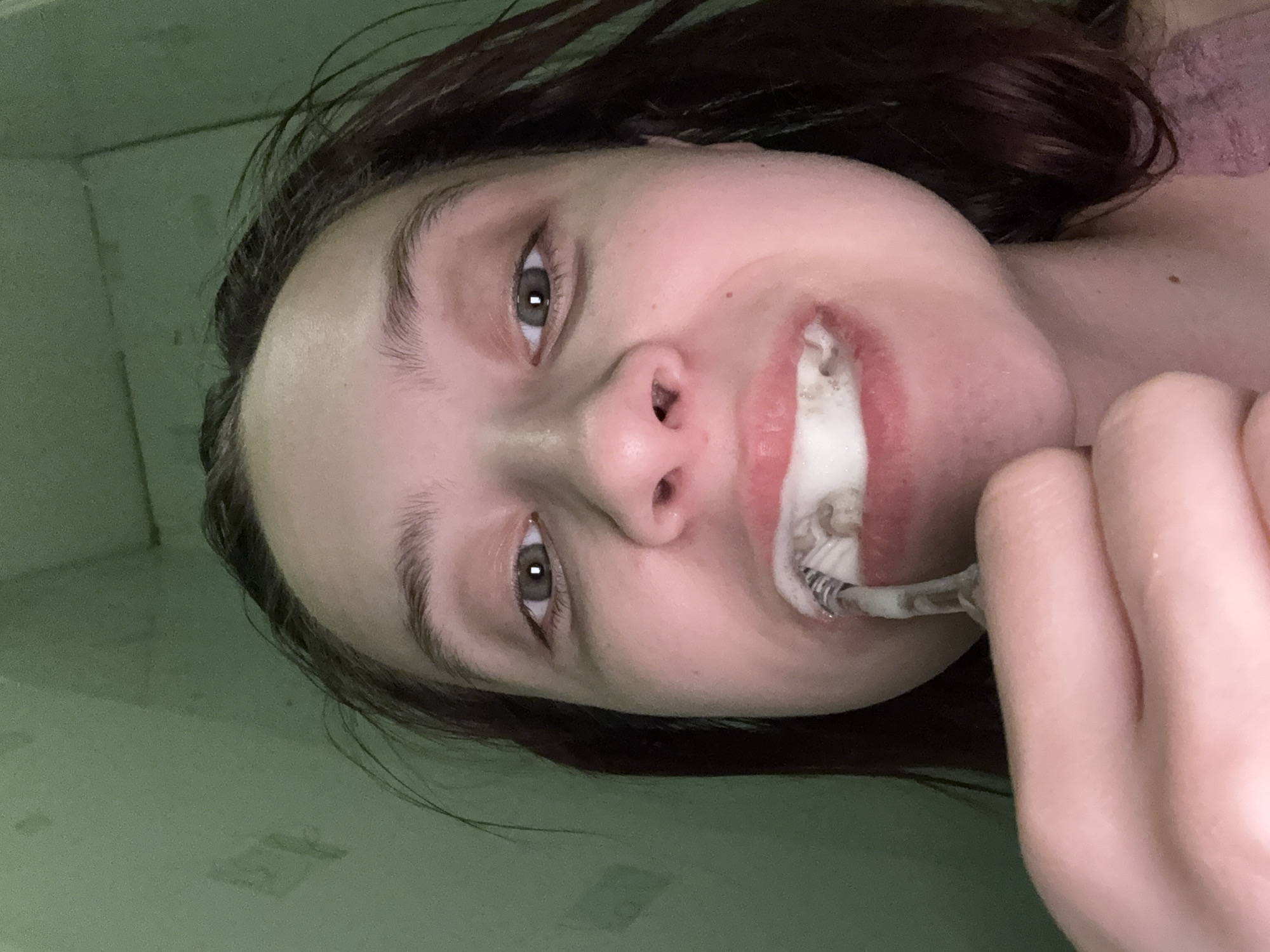 A close-up, angled shot depicts a woman brushing her teeth. She holds the toothbrush to her mouth with her right hand. The bathroom wall is a light green. It is not possible to verify the location within the city of Прикамский, Russia from the provided image.