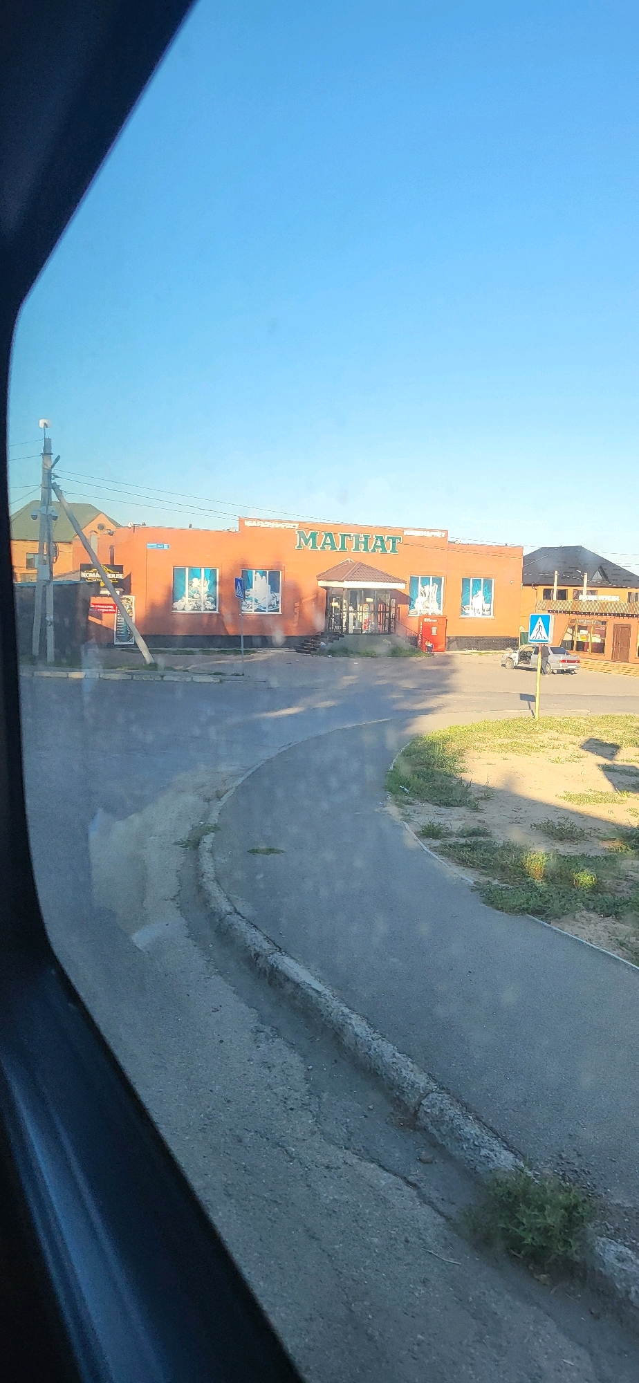 The image shows a street scene in Oskemen, Kazakhstan. The prominent feature is a single-story, orange-colored building with a green sign that reads "МАГНИТ" (Magnit). The building appears to be a retail store, possibly a supermarket, with several windows and an entrance with a covered canopy. A red vending machine is visible next to the entrance.
In front of the building, a paved road curves. There are several parked vehicles, including a small ATV and a silver sedan. Blue and white directional signs are present on poles along the road. A concrete curb separates the road from a grassy area with some weeds.
A utility pole with electrical wires stands to the left of the building. The sky is clear and blue. The overall impression is of a typical street in a residential or commercial area of a city. The image seems to be taken from inside a vehicle, as suggested by the dark frame on the left side, possibly a car window.