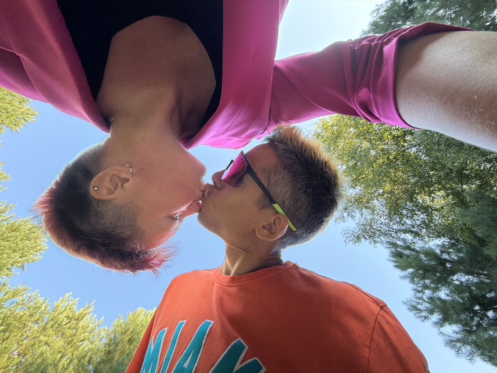 In an outdoor setting with a clear blue sky and lush green trees, two individuals are captured in an intimate moment. The image is taken from a low-angle perspective, looking upwards.
One individual, with short hair dyed with pinkish-red tones, is wearing a vibrant fuchsia jacket. They are leaning in for a kiss, their face partially obscured by the jacket and their head tilted downwards. An earring is visible on their left ear.
The other individual, facing the camera, has short, multi-toned hair and is wearing pink-tinted sunglasses. They are dressed in a bright orange t-shirt with a graphic print that appears to say "MIAMI" in teal and white letters. Their lips are pressed against the other person's.
The scene suggests a warm, sunny day, possibly in a park or garden. The specific location in the city of Chaikovsky, Russia, is not discernible from the visual information provided in the image itself.