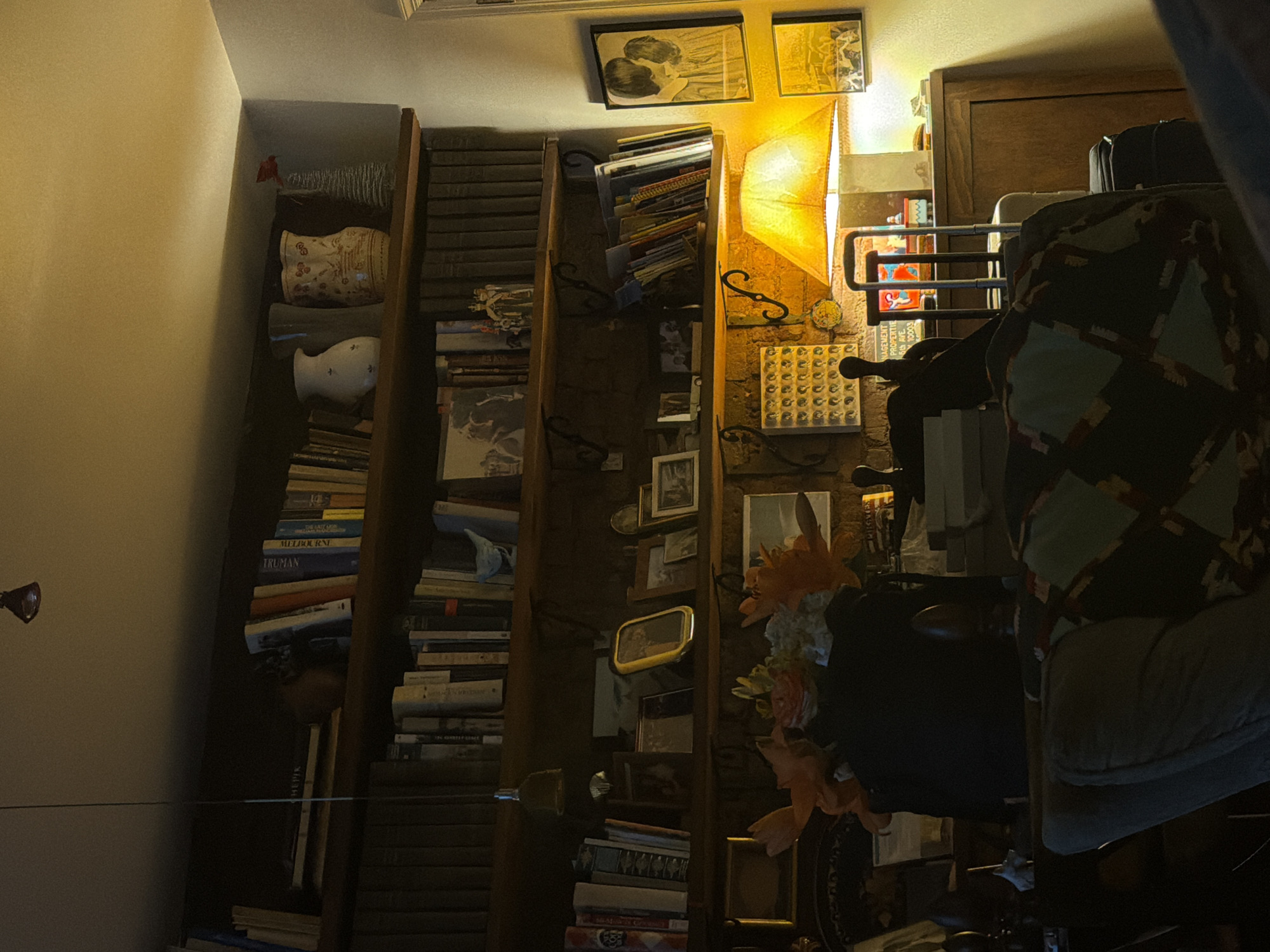 The image shows an interior scene, likely a home, with a focus on a bookshelf and nearby furniture. The bookshelf is filled with books, arranged vertically, and various decorative objects. Adjacent to the bookshelf, there is a lamp emitting a warm, yellow light. Various pictures and frames adorn the wall space around the bookshelf and lamp. In the foreground, there is a chair with a patterned covering. Based solely on the visual information, the location cannot be determined to be in New York City. The scene presents a cluttered but cozy domestic environment.
