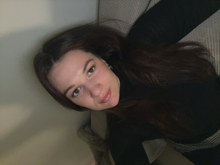 A fair-skinned woman with auburn hair is angled towards the viewer. She is wearing a black turtleneck. Her hair is parted in the middle, and she has light brown eyes and pink lips. Part of a light-colored surface is visible to her left, possibly a wall or sofa. Part of what looks like a beige garment with a furry trim is seen to the lower-left of her neck. The lighting appears soft and diffused.