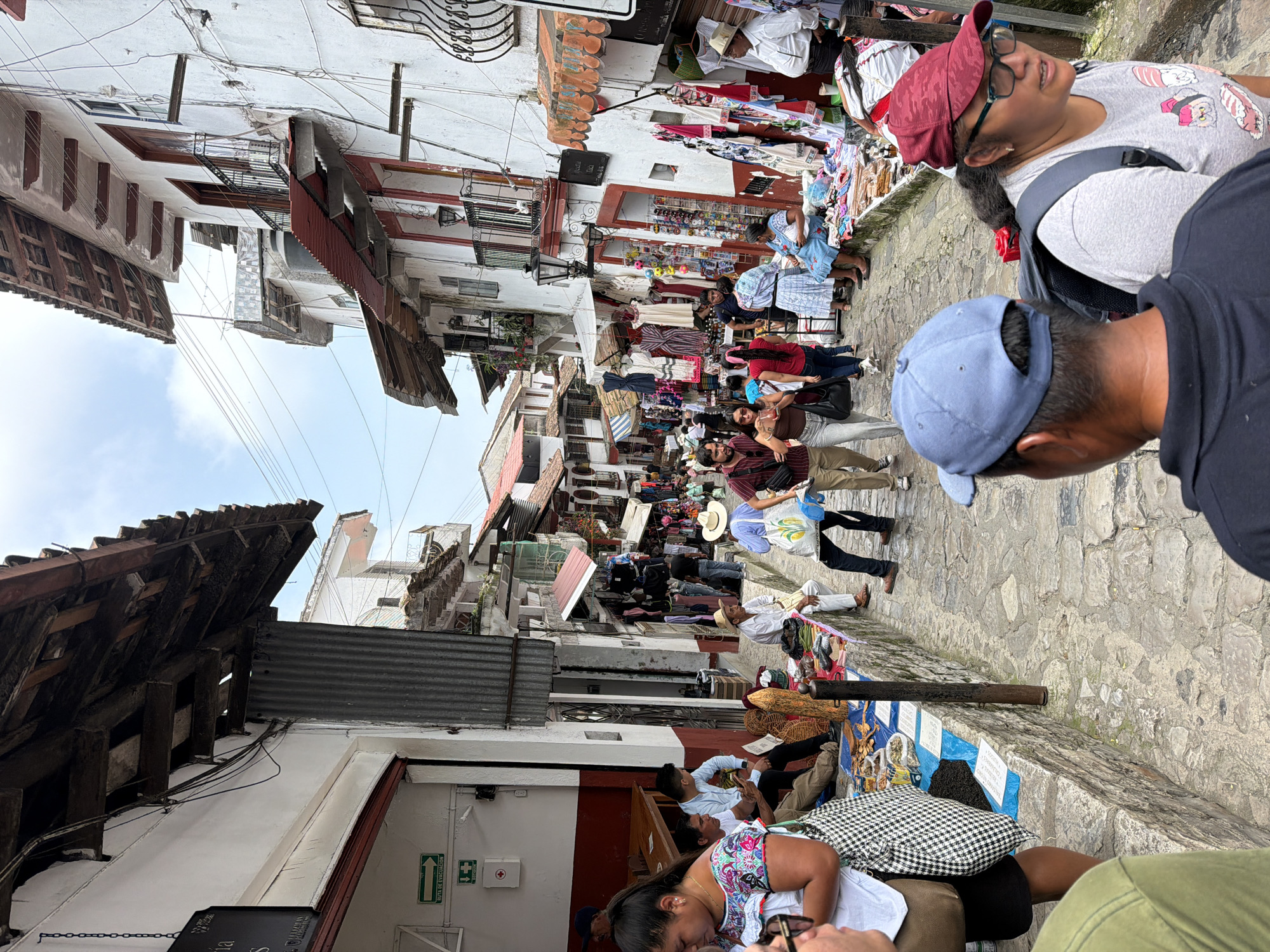 This image captures a bustling street scene in Cuetzalan, Mexico. The narrow, cobblestone street is lined with whitewashed buildings, some adorned with red-tiled roofs. Vendors are selling merchandise, including clothing, jewelry, and handicrafts, that spread out onto the street. Many people walk through the street, including tourists and locals wearing casual clothes. The sky is visible through the gaps between the buildings, appearing partly cloudy. A few individuals are visible in the foreground, including one wearing a red cap and another wearing a blue cap.
