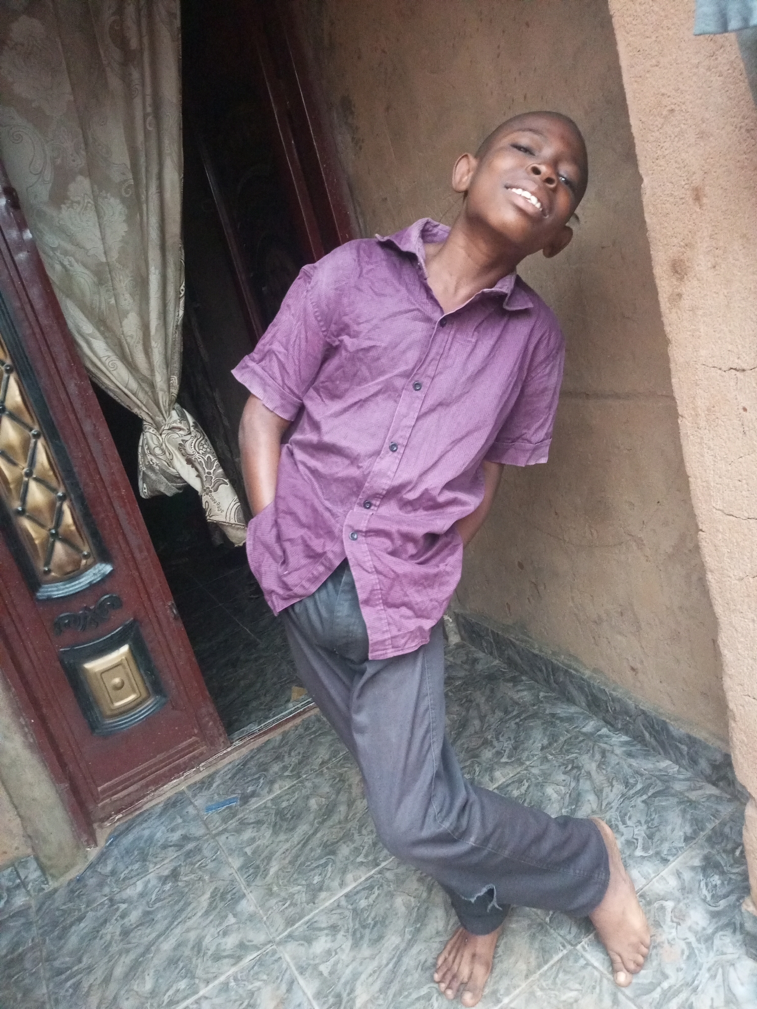 A young Black male, appearing to be in his early teens, is pictured standing outside a building with his left hand in his pocket and his right arm extended backward. He is wearing a short-sleeved, purple collared shirt and dark gray pants. The background includes a brown textured wall, a closed wooden door with decorative metal accents, and a beige curtain. The ground is tiled with a marbled pattern. The scene appears to be located in an outdoor area, possibly an alley or entryway, consistent with a residential setting in Tempure, Nigeria.