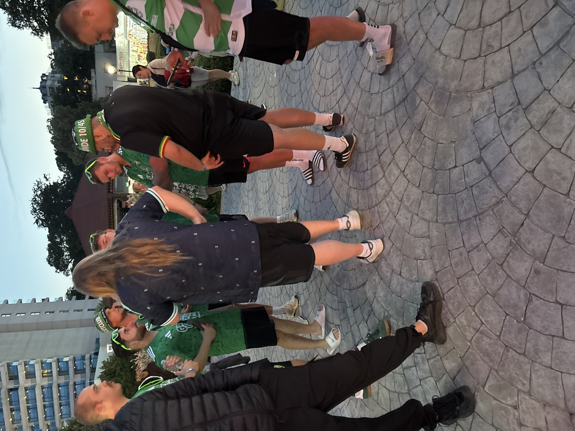 The image depicts an outdoor social gathering at dusk or early evening in Varna, Bulgaria, specifically identifiable by a prominent modern, multi-story white building with balconies in the background, characteristic of coastal developments, alongside an older building partially obscured by trees and distant illuminated market stalls. The ground is paved with a gray, circularly patterned stone.
Approximately ten individuals, primarily adult males, are present, standing in small clusters and engaged in conversation. Several men are wearing green sports-themed apparel: green t-shirts, green sunglasses, a green bucket hat with "SPORTCLUB" text, and another green bucket hat with "UNDE 10/45" text and Rasta-colored banding. A man on the far right wears a white jersey with green horizontal stripes. A man on the far left wears a black puffer jacket and dark pants, possessing a mustache. Another man in the center foreground, facing away, has long brown hair and wears a dark blue patterned shirt and black shorts. In the mid-ground right, a woman in a light jacket and dark shorts is observed holding a smartphone. Footwear varies and includes black sneakers, white Nike slides, Adidas slides, and Adidas sneakers (black with white stripes), often worn with white socks. One man holds a clear plastic bottle. The scene is lit by the ambient twilight and artificial lights from the background structures.