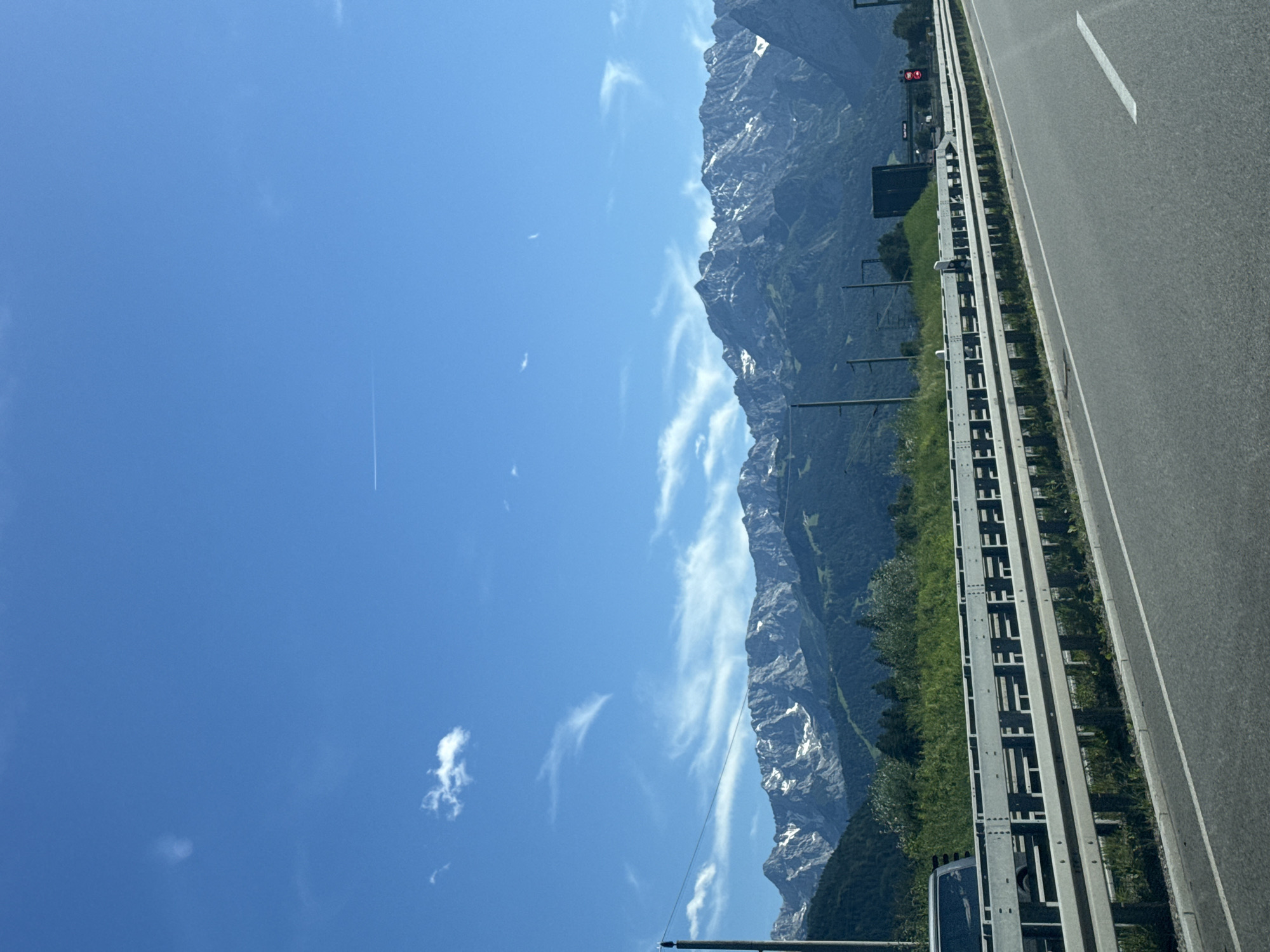 The image depicts a scenic view from a highway in Oberau, Germany. The foreground shows a grey asphalt road with white lane markings, bordered by a metal guardrail and a grassy median. Beyond the highway, a dense line of green trees and vegetation stretches across the landscape, leading to a majestic mountain range in the background. The mountains are rugged and imposing, with some peaks still capped with snow. The sky is a vibrant blue, with a few wispy white clouds scattered across it, and a contrail from an airplane is visible. The overall impression is one of natural beauty and vastness, with the highway serving as a modern element cutting through the pristine environment.