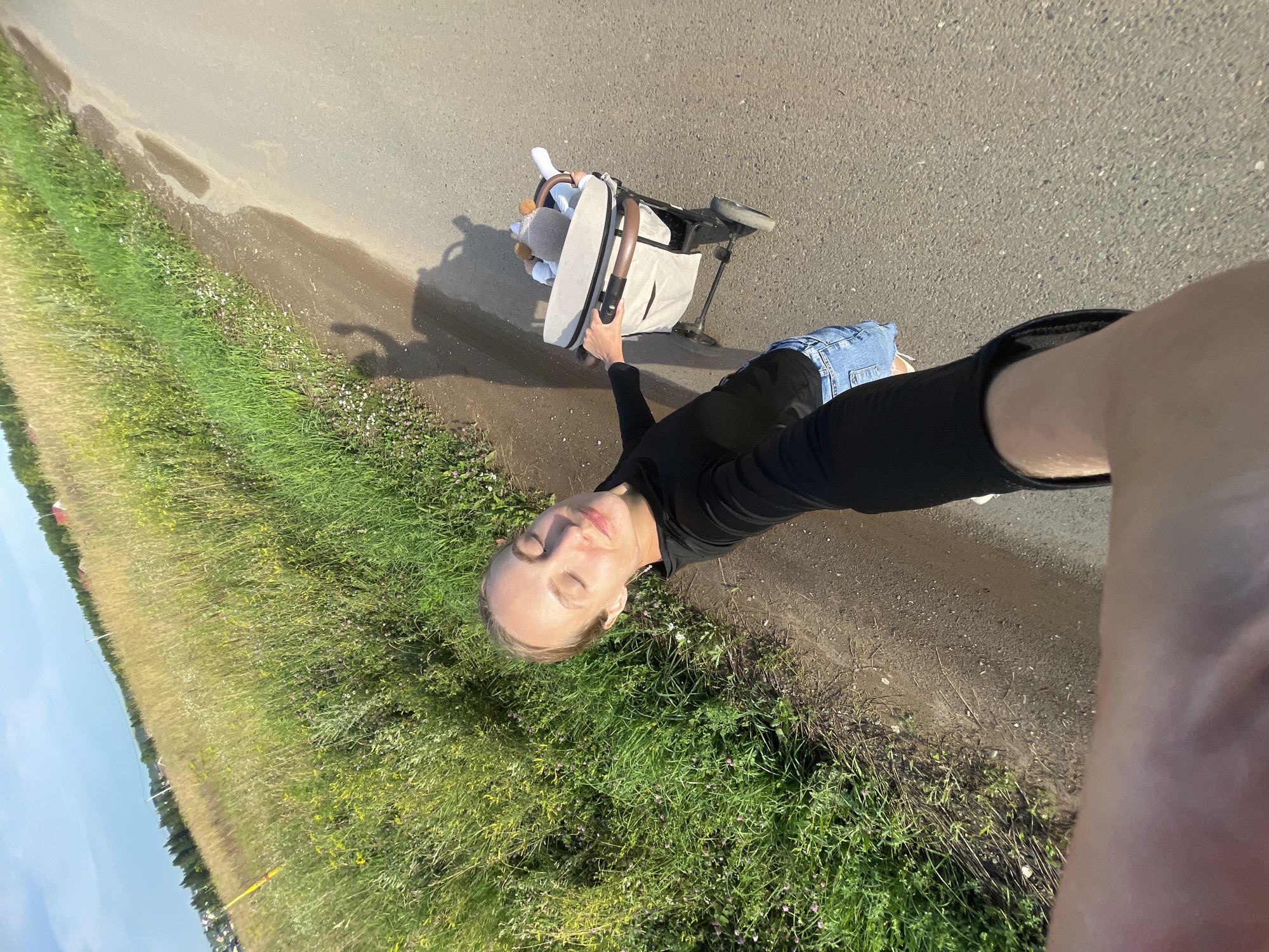 In an overhead, selfie-style shot, a person with closed eyes and a serene expression, positioned in the foreground, pushes a light-colored baby stroller along a paved path. The stroller contains a baby or stuffed toy. The person wears a black long-sleeved shirt and denim shorts. To the left of the path, a field of tall green and yellow grass stretches towards a distant tree line and scattered buildings, indicative of the rural outskirts of Podshivalovo, Russia. The sky above is a pale blue with some wispy clouds. The lighting suggests it is late afternoon, casting long shadows.