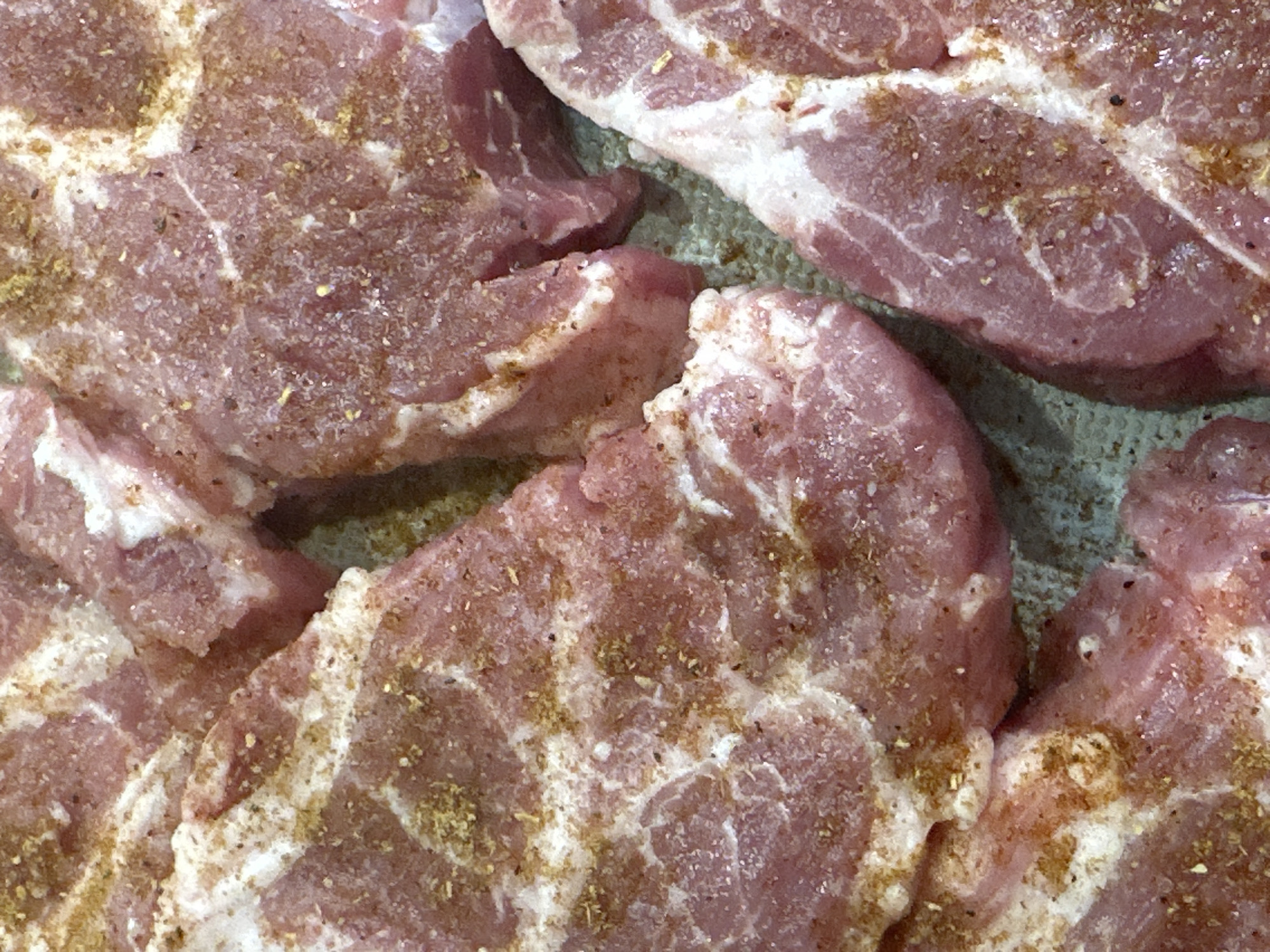 The image depicts several pieces of raw pork, likely pork chops or cutlets, seasoned with a brownish spice blend. The meat is pink with marbling of white fat. The background is a textured surface, possibly a plate or cutting board. No people are visible in the image. The location is unknown.