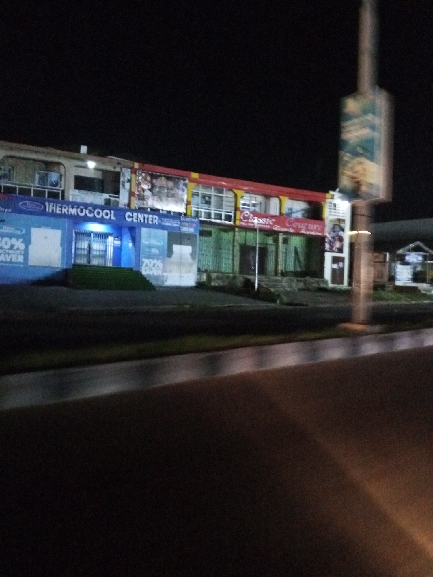 The image depicts a nighttime scene in Lokoja, Nigeria, featuring a row of commercial buildings along a street. The most prominent establishment is "THERMOCOOL CENTER," identified by a blue facade and illuminated signage. Adjacent to it, another business labeled "Classic Couture" is visible, with green-colored walls and signage. The buildings appear to be two stories tall, with windows and doors indicative of commercial spaces. A tall, vertical billboard stands to the right of the buildings, partially illuminated. The foreground shows a street or roadway with a blurred appearance, suggesting movement, possibly from a vehicle. The overall environment is dimly lit, characteristic of a night scene in an urban setting. There are no individuals clearly visible in the image.