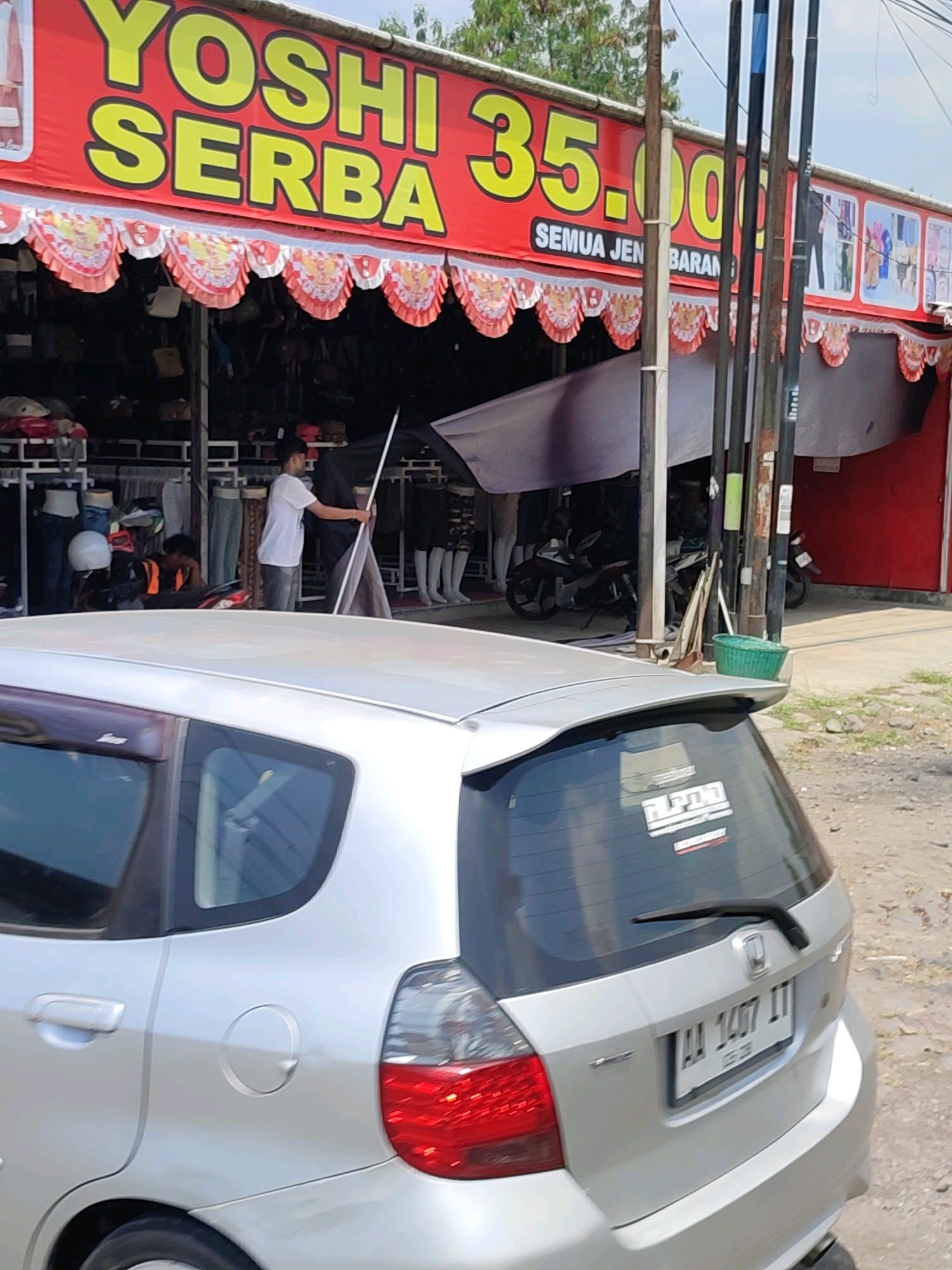 The image shows a street scene in Tanggulrejo, Indonesia, with a shop named "YOSHI SERBA 35.000" prominently displayed on a red banner. The shop appears to be selling various items, with clothing and bags visible on display racks. A silver hatchback car, likely a Honda Jazz based on the rear design, is parked in front of the shop. Two men are present: one in a white t-shirt and grey pants is standing inside the shop, holding a long pole and appearing to adjust a tarp or display. The other man, wearing a black cap and an orange vest over a black shirt, is seated on a motorcycle within the shop's premises, looking down. The environment suggests a warm, possibly humid climate, with a clear sky and some greenery in the background. The shop has a festive facade with red and white scalloped trim. A green basket is visible near some vertical poles outside the shop. The license plate of the car is visible as "AA 1407 IT".