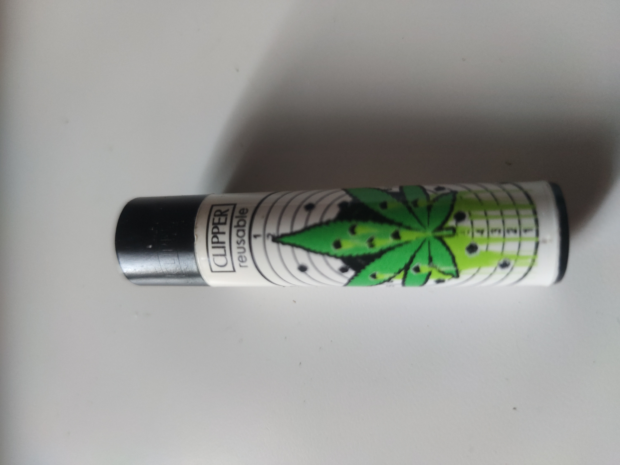 The image shows a Clipper brand reusable lighter lying horizontally on a white surface. The lighter has a black top and a white body. The body is decorated with a green marijuana leaf design and has numbers 1 through 5 marked along its length. "CLIPPER" and "reusable" are printed on the side near the black top. The location is not provided.