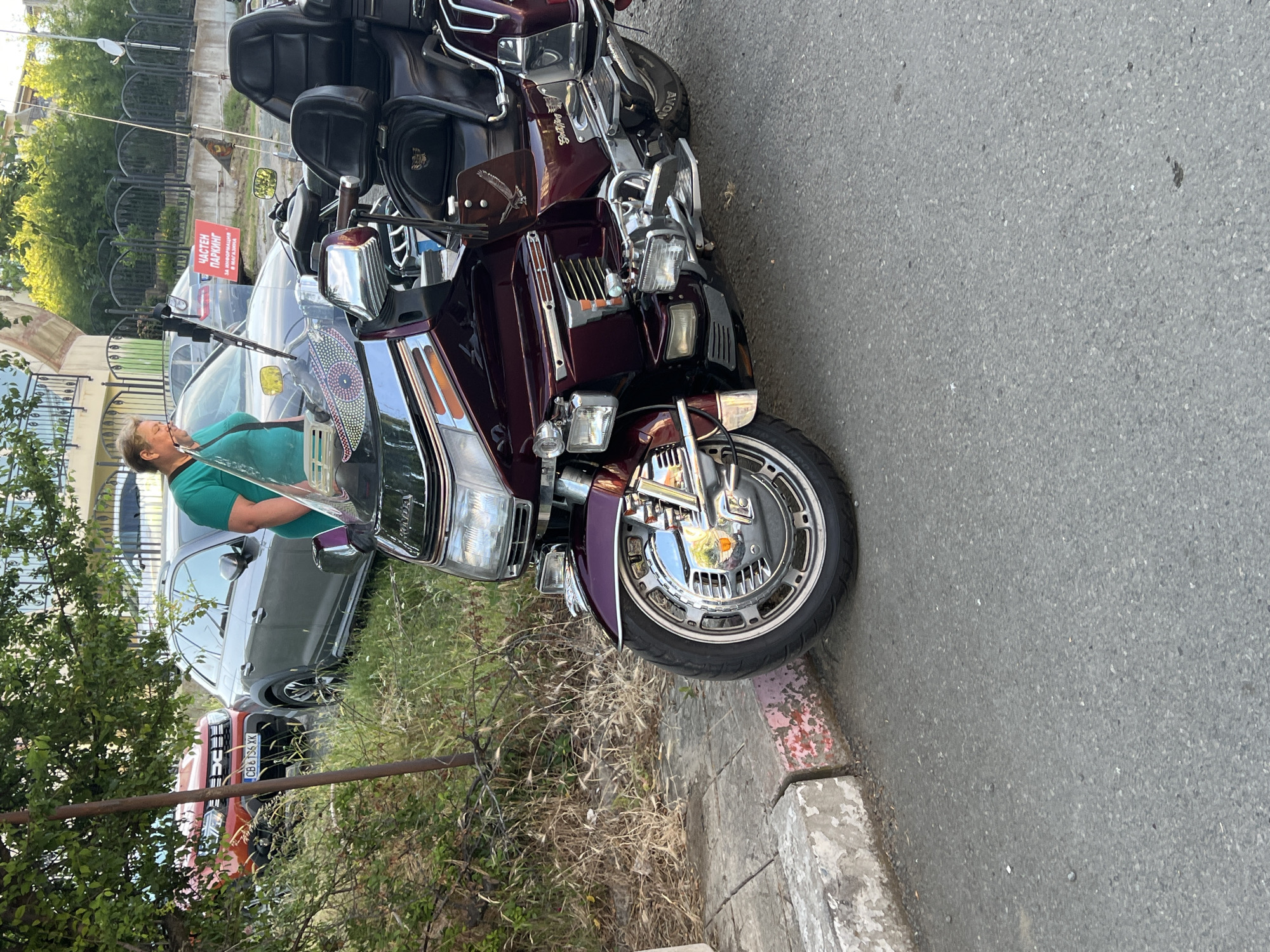 The image shows a close-up of a maroon Honda Gold Wing motorcycle parked on an asphalt road. The motorcycle is adorned with chrome accents, including its wheels, engine components, and handlebars. The front wheel features a prominent chrome rim with intricate detailing, and a circular chrome cover in the center. The motorcycle's plush leather seats are visible, as well as a windshield with a decorative decal.
In the background, a woman in a teal top is partially visible, leaning over the motorcycle. Behind her, two cars are parked: a grey Audi SUV and a bright orange Dacia Duster with a Bulgarian license plate reading "CB 136 XK." The scene appears to be in a semi-urban or residential area, with overgrown vegetation and a concrete curb running along the edge of the road. A red sign with white lettering, which reads "ЧАСТЕН ПАРКИНГ" (Private Parking), is also visible in the background. This suggests the location might be near a commercial establishment or private property. The overall lighting indicates it is daytime, with soft sunlight casting gentle shadows.