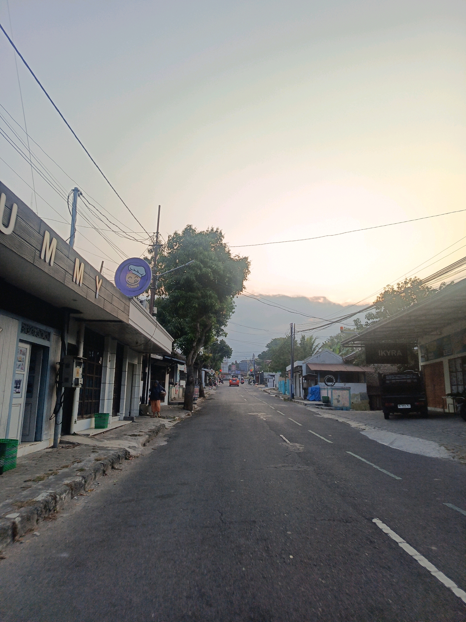 The image depicts a street scene in Candirejo, Indonesia, likely during the late afternoon or early morning due to the soft, diffused light. The street is paved and has white lane markings. On the left side of the road, there is a building with the sign "YUMMY" in large letters. Several shopfronts are visible, some with open doors. A person wearing an orange skirt and dark top is standing on the sidewalk in front of the "YUMMY" building, carrying a basket. Green trash bins are placed near the shops. A circular sign with a cartoon chef is mounted above the "YUMMY" entrance.
Further down the street, trees line the right side of the road, casting shadows. More buildings and shopfronts are visible, including one with the sign "IKYRA." A dark-colored pickup truck is parked on the right side of the road. In the distance, several vehicles, including a red car, are visible on the street. The sky is bright with a gradient of light, suggesting either sunrise or sunset, with some clouds visible towards the horizon. Overhead, numerous electrical wires crisscross the scene, suspended from poles.