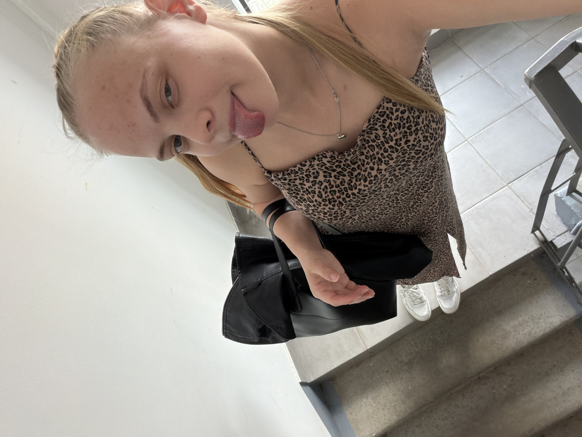 A young woman takes a selfie while standing on a concrete staircase. She has blonde hair, light skin, and is wearing a leopard-print dress and white sneakers. She is sticking her tongue out. She is also wearing a necklace. She carries a black leather purse. The background shows a white wall and grey staircase. It is not possible to determine if the location is Izhevsk, Russia, based on the image alone.