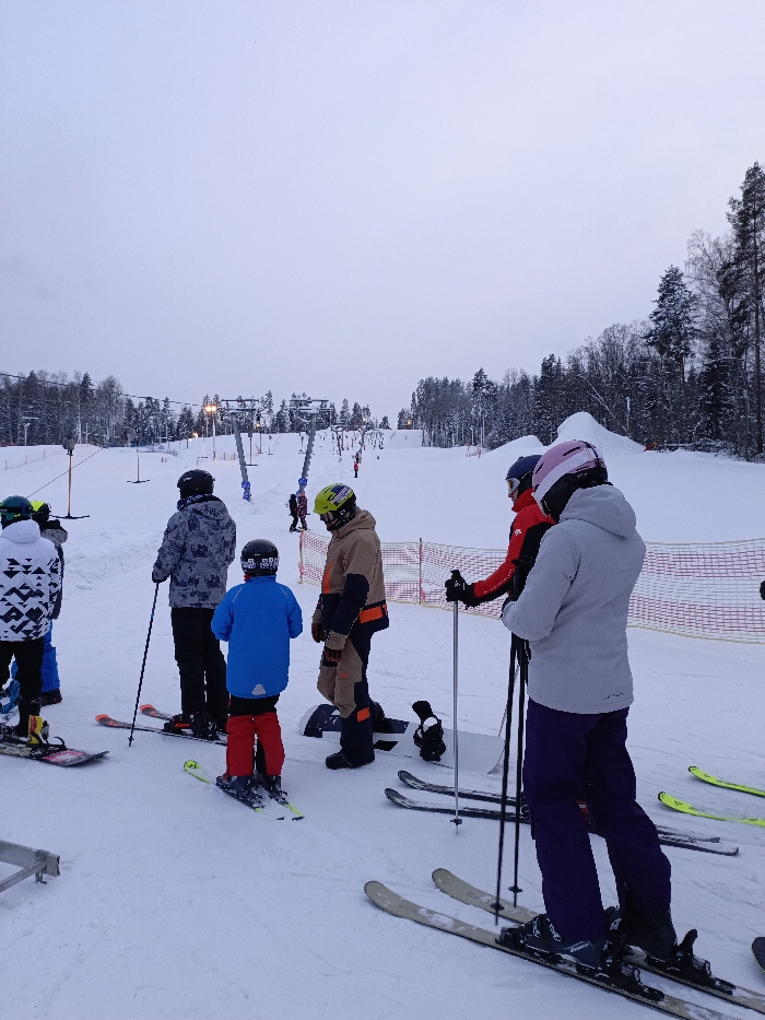 This image captures a group of people at the base of a ski slope, likely at a ski resort, on an overcast winter day. The sky is a uniform, light grey, suggesting either early morning, late afternoon, or consistently cloudy weather, with no direct sunlight visible.
The ground is entirely covered in white snow, indicating a cold, wintry environment. In the background, snow-covered evergreen trees line the hills, characteristic of a mountainous or hilly ski area.
Several ski lift pylons, appearing to be for T-bar or button lifts, ascend the slope in the mid-ground. Lights are visible on some of these pylons, suggesting the area might be lit for night skiing or for improved visibility during low-light conditions. Orange safety netting runs along the sides of the slope, separating areas or indicating boundaries. Other skiers and snowboarders can be seen further up the slope near the lift.
In the foreground, a group of approximately six individuals are gathered. They are all dressed in full winter sports gear, including helmets, ski jackets, and ski pants, indicating they are prepared for skiing or snowboarding. Most are wearing skis, while at least two individuals are on snowboards.
From left to right:
* A person in a white and black patterned jacket and black helmet stands on a snowboard, facing away from the camera.
* Next to them, a person in a grey and white patterned jacket and black helmet stands on skis, holding ski poles, also facing away.
* A younger individual, likely a child or teenager, in a bright blue jacket, red pants, and black helmet, is on skis and facing away.
* An adult in a tan and black ski/snowboard suit with orange accents and a green helmet stands on a snowboard, looking to the left.
* Further to the right, two people are standing on skis, holding poles. One wears a red jacket and dark helmet, while the other, slightly in front, wears a light grey jacket, dark purple pants, and a pink helmet. Both are facing to the right.
* A pair of skis and ski poles are partially visible on the far right, suggesting another person just out of frame.
The group appears to be waiting in line for the ski lift or perhaps regrouping at the bottom of a run. The atmosphere seems calm, typical of people enjoying a winter sports outing. No discernible text is visible anywhere in the image.