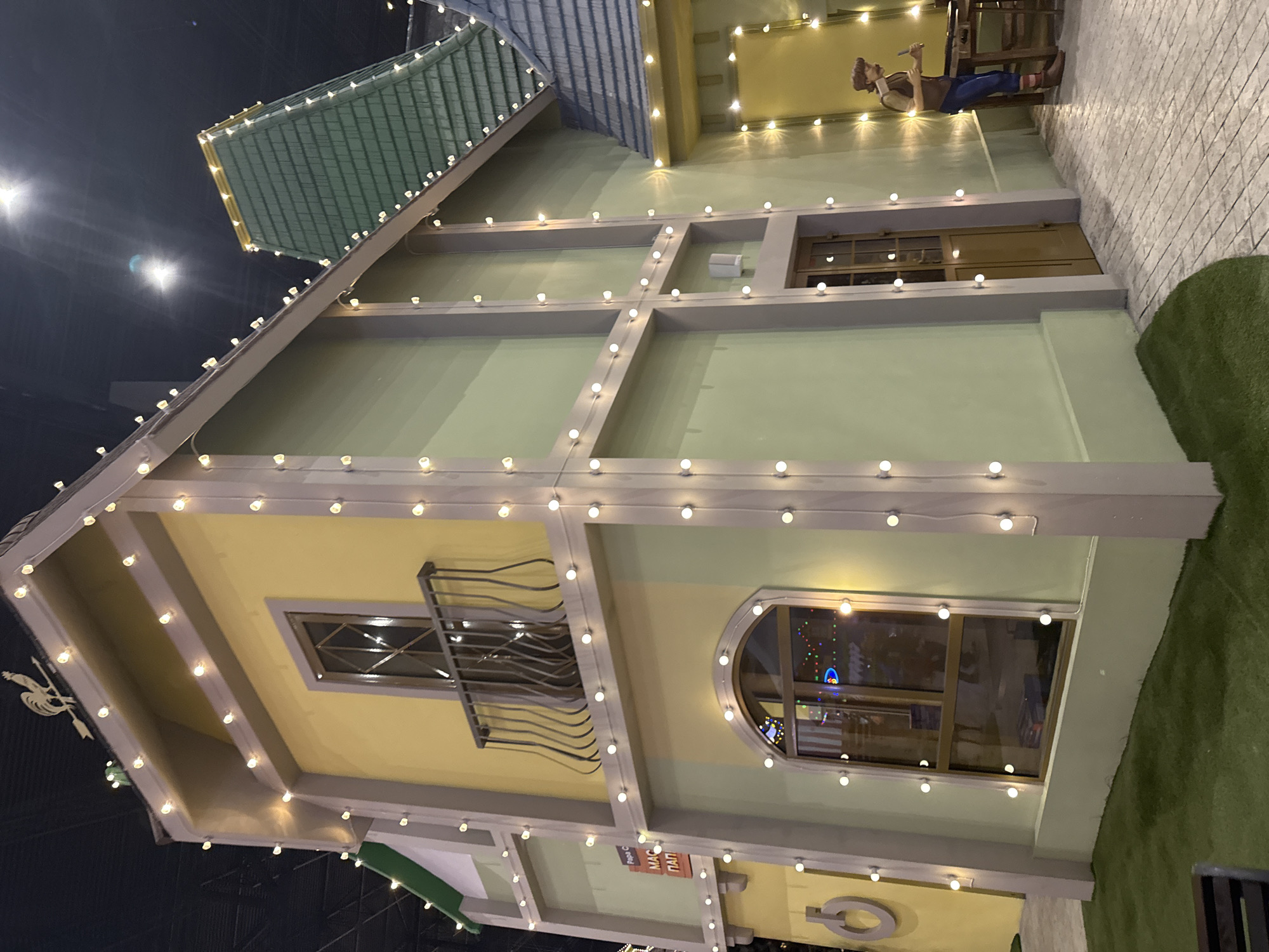 The image depicts a whimsical, illuminated building exterior, festooned with strings of white lights. The building has a sloping roof with what appears to be shingles and is painted in muted shades of yellow and green. It features decorative architectural elements, including a window with a wrought-iron balcony and an arched window. There are signs in Cyrillic script, indicating the location may be in Russia. At the top of the building is a figure sitting at a table, implying this might be a fanciful structure for an event or display. The scene is set against a dark backdrop, emphasizing the illuminated building and creating a dreamlike atmosphere. Based on the signs visible "МАС ПАП", the location is most likely within Moscow, Russia.