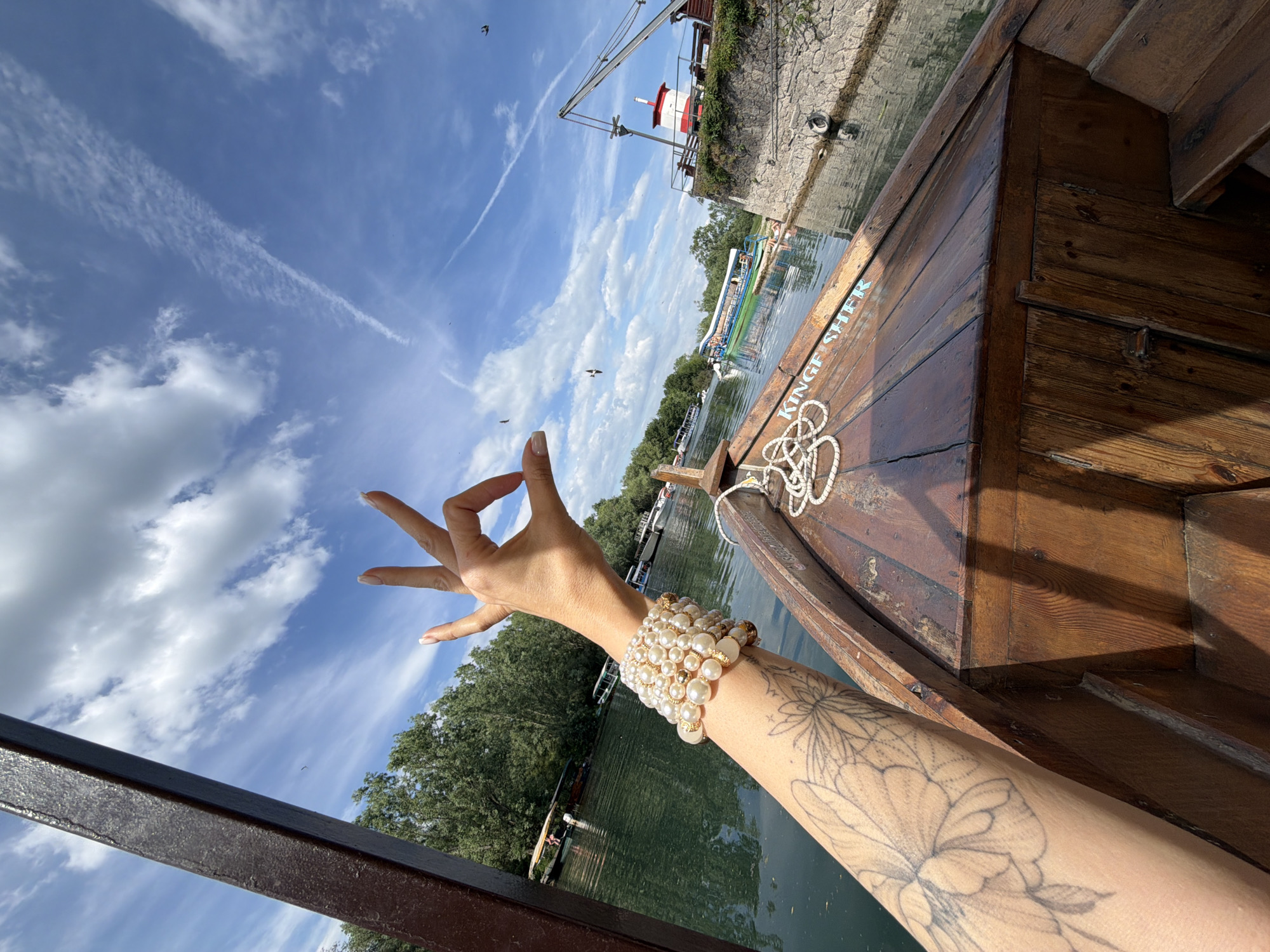 A low-angle shot shows a woman's arm outstretched in front of a boat, with a partially visible tattoo, a pearl-like beaded bracelet on the wrist, and a hand performing a mudra against a background of sky with light white clouds. The boat is wooden with visible grains and rope details. In the background are buildings along the water's edge and lush green vegetation, suggesting the location of Boljevići, Montenegro, given the visible landmarks in the full view. The camera is held at an angle, creating a dynamic composition. Birds are flying above the trees, and additional boats can be seen in the distance.