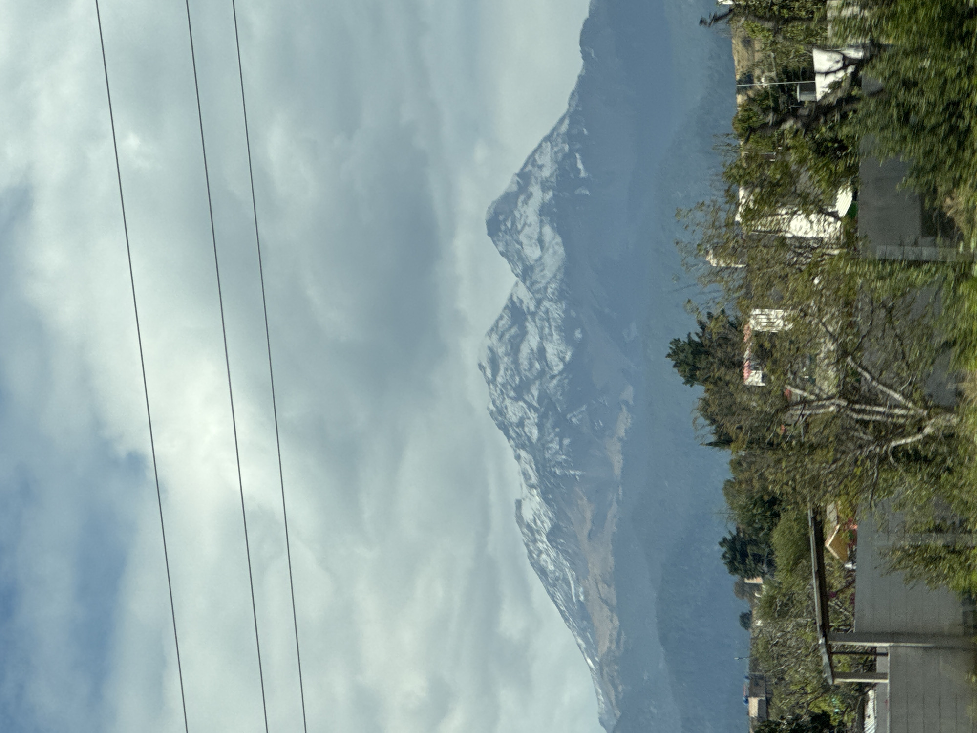 A slightly blurry, rotated view shows electrical wires against a cloudy sky and the snow-dusted peak of La Malinche mountain, as seen from Santa Rita Tlahuapan, Mexico. Adjacent buildings and trees are visible on the right side of the frame.
