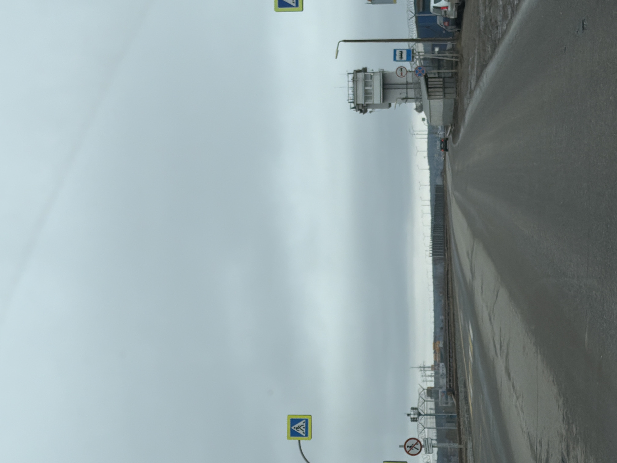The image shows a wet road in the city of Chaikovsky, Russia. The sky is overcast. There are road signs indicating a pedestrian crossing and no pedestrian crossing. A complex structure possibly related to border control is visible on the left side of the road. A row of buildings/infrastructure is visible in the distance along the left side of the road. Cars are parked in the distance on the left.