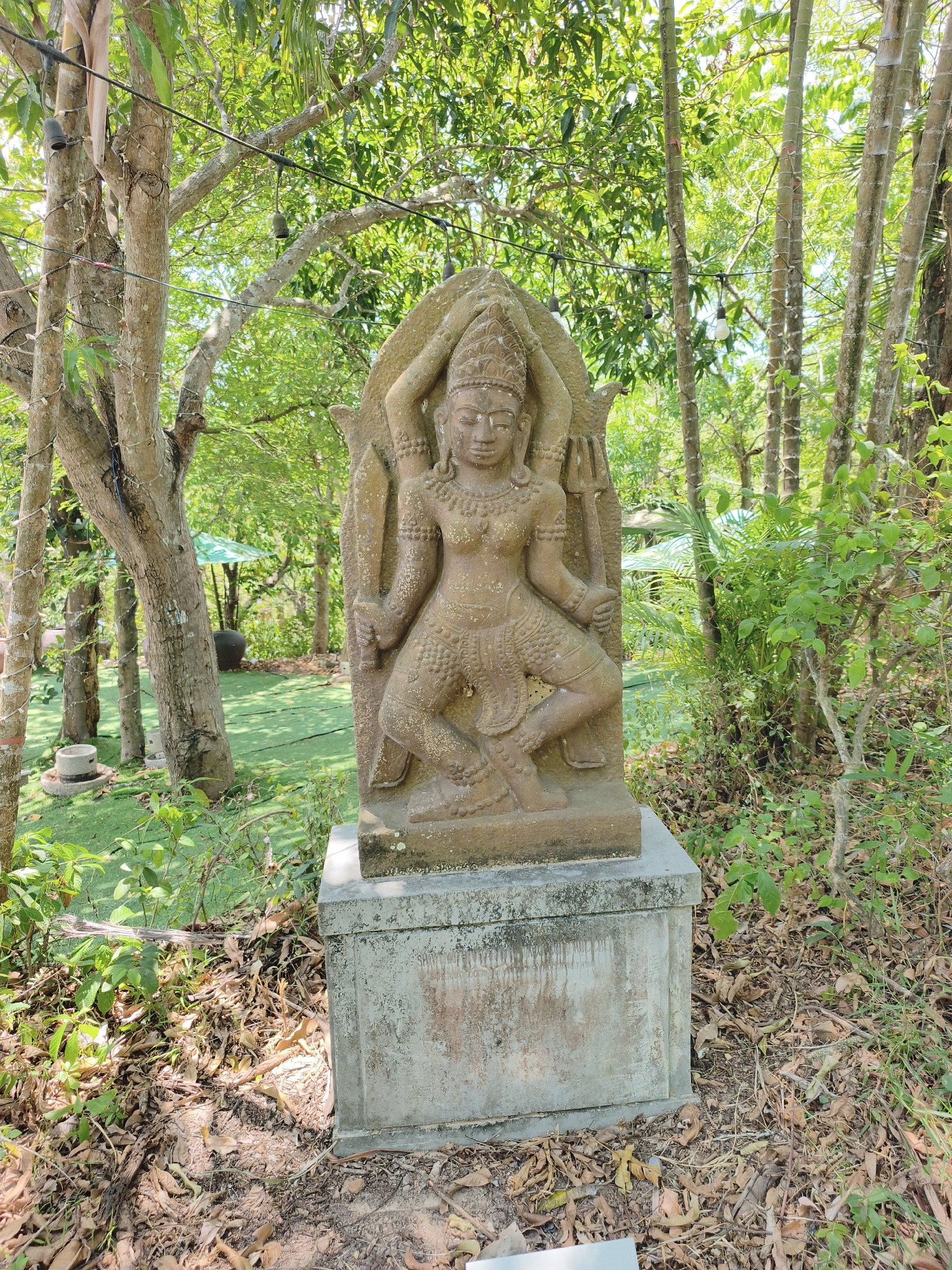 The image displays an ancient stone statue, possibly of a deity, situated outdoors on a concrete pedestal. The statue is intricately carved, depicting a figure with multiple arms, adorned with elaborate jewelry and headdress. The statue is positioned in a lush, green environment with numerous trees and foliage surrounding it. In the background, a green umbrella shade and a large dark pot are visible, suggesting a garden or outdoor seating area. The ground around the statue is covered with dry leaves and twigs. The lighting indicates it is daytime, with sunlight filtering through the trees. The overall scene suggests a tranquil and possibly historical or religious site within a natural setting. There are no people present in the image, and the focus is solely on the statue and its surroundings. The statue is located at the Po Nagar Cham Towers in Tuy Hoa, Phu Yen Province, Vietnam.