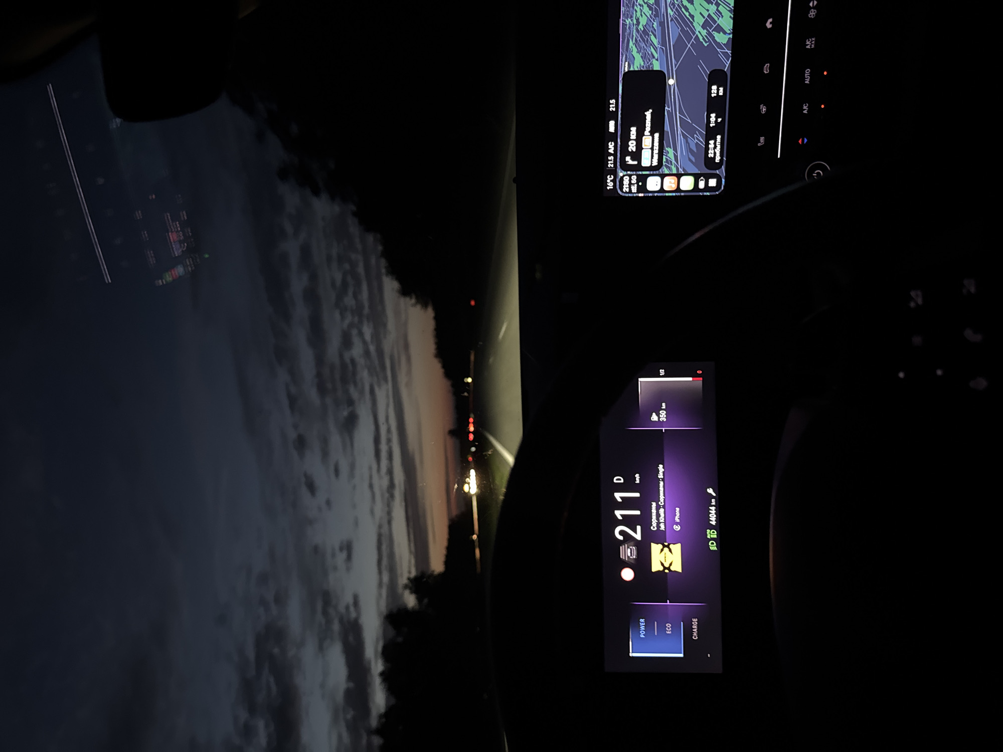 The image depicts the interior of a car driving at night. The speedometer indicates a speed of 211 km/h. The music playing is "Сюрикены" by Jah Khalib on iPhone. A navigation system shows a route with approximately 20 km remaining to Poznań, near Warszawa. The car's air conditioning is on, set to 21.5°C. The time is 2:50, with an estimated arrival time of 22:54. The car has 350 km left. The driving mode is set to Eco. The odometer reads 44044 km. Through the windshield, a road stretches into the distance with headlights of oncoming vehicles. The sky above is dusky.
