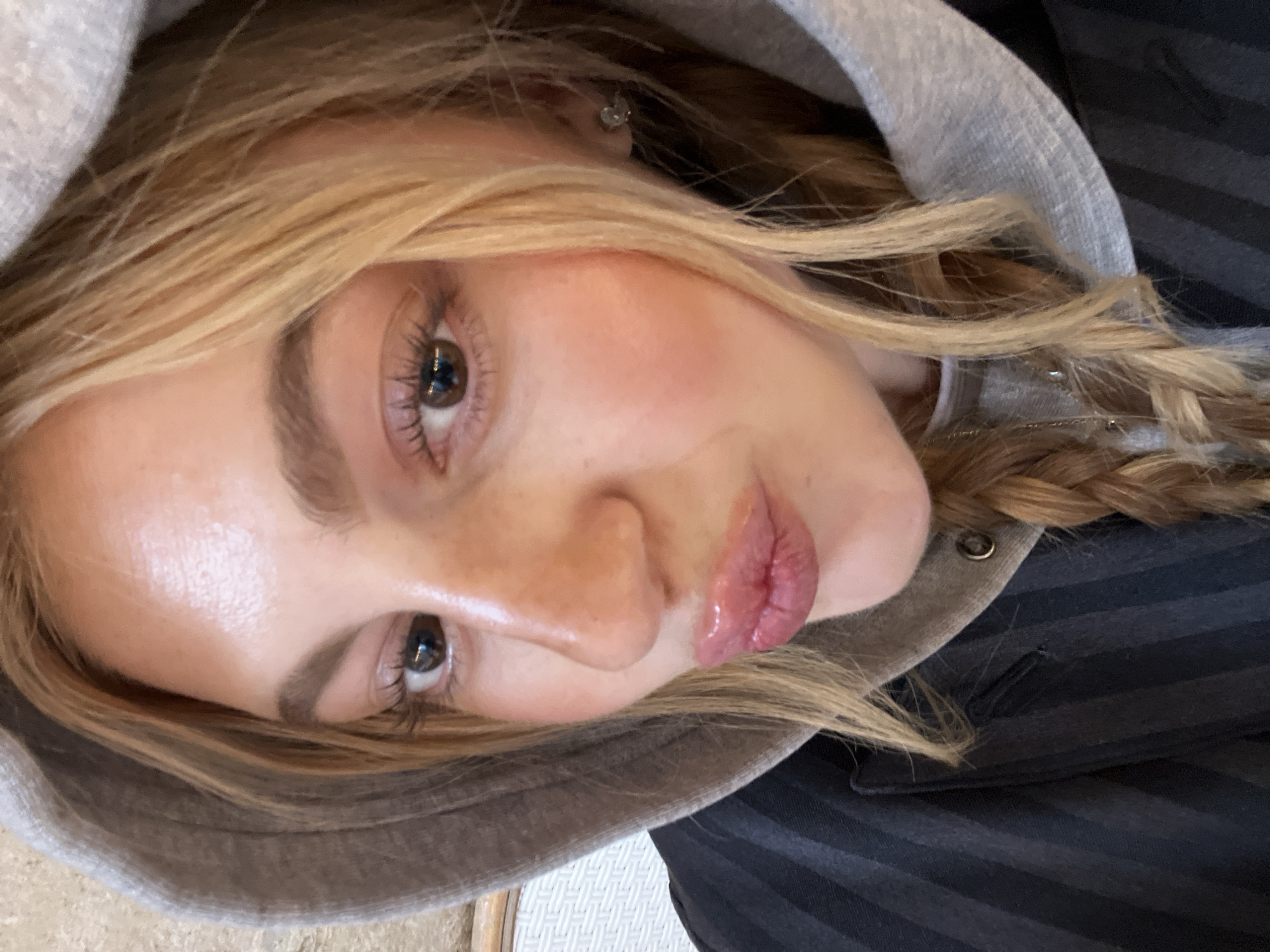 A young woman with light brown hair is wearing a gray hooded sweatshirt over a dark striped garment. She is facing the camera with a slight pout. Her skin tone appears fair. She has brown eyes, and her eyebrows are lightly filled in. Her lips are slightly glossy. She has one braid in her hair. An earring is visible in her left ear. The background is blurry, but the edge of what appears to be a white or light-colored surface is visible. There is no information in the image to indicate the location is Paris, France.