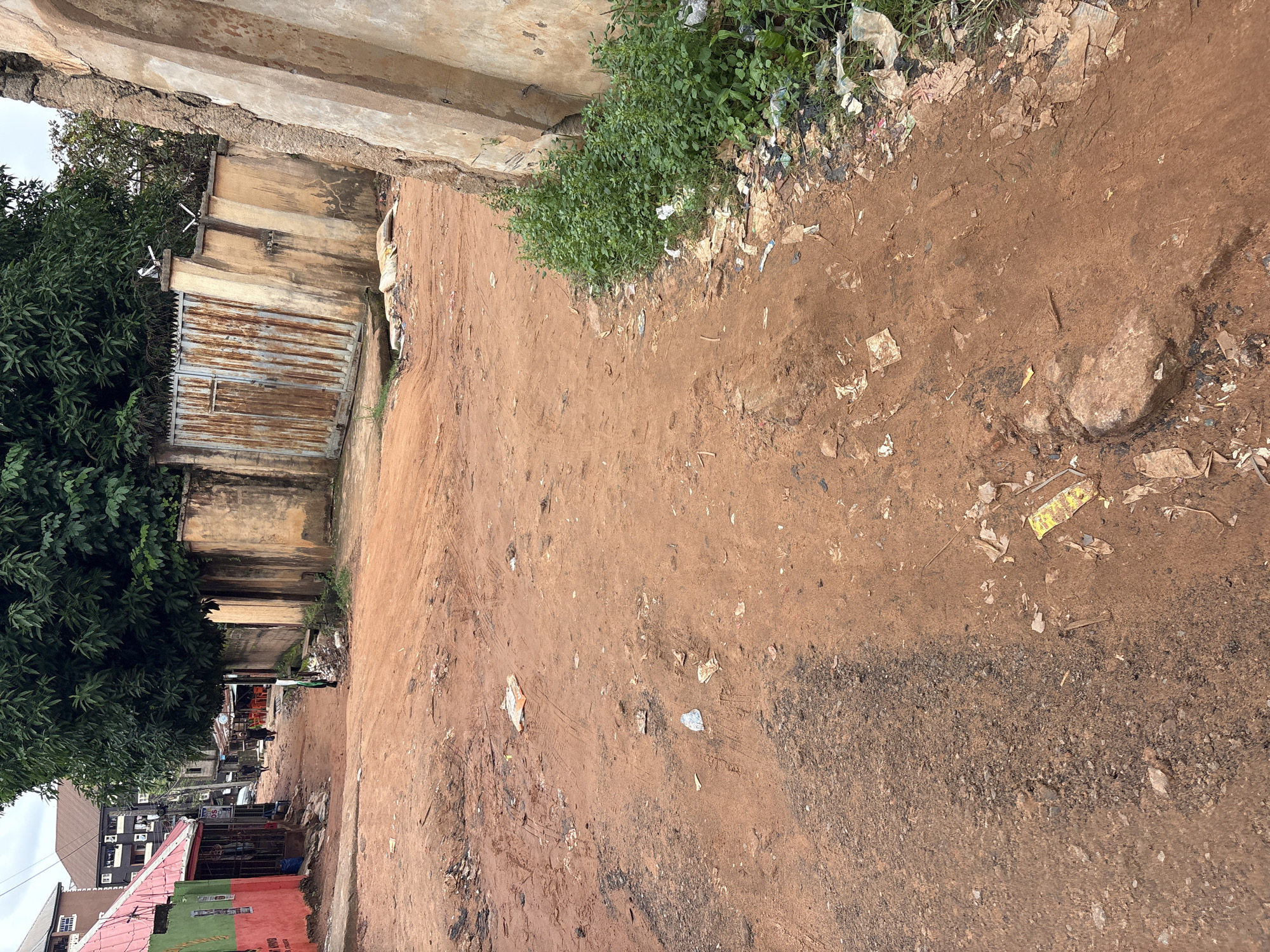 The image depicts a dirt road in Nkpor, Nigeria. The road is unpaved and appears to be muddy in places, indicating recent rainfall. The surrounding environment is a mix of residential and commercial structures. On the left side of the road, there is a building with a red and green facade, possibly a shop or business. Further down the road, more buildings can be seen, including a multi-story structure. A large tree provides shade over a portion of the road. On the right side of the road, there is a rusty metal gate and a weathered concrete wall. The ground is littered with debris, including plastic bags, wrappers, and other trash, which is also accumulating along the edge of the wall. There are a few people visible in the distance, walking along the road. One person is walking towards the camera, while others are further down the road, appearing to be engaged in daily activities. The overall scene suggests a bustling, yet unkempt, urban environment common in many parts of Nigeria.