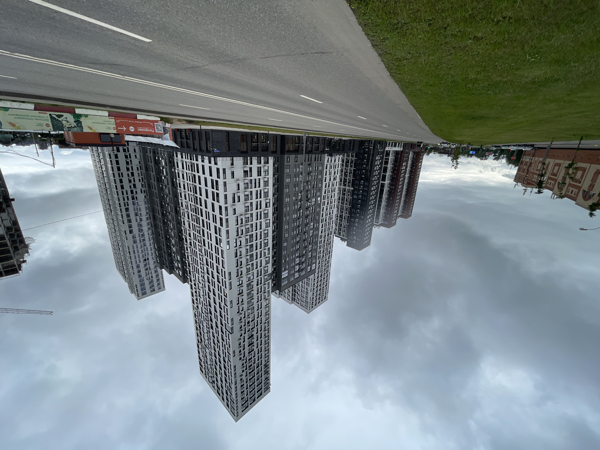 The image shows a wide, empty asphalt road in a modern urban environment under a cloudy sky. On the right side of the road, a series of tall, modern residential buildings with white and dark gray facades and numerous windows are lined up. The buildings are of varying heights and architectural styles, suggesting a new development. A construction crane is visible in the distance to the right, indicating ongoing construction. On the left side of the road, there is a strip of green grass, followed by a multi-story building with a reddish-brown facade and arched windows, appearing to be older than the residential towers. Further down the road on the left, more cars are visible, and trees are planted along the sidewalk. In the foreground, the road is divided by white lines, and the grass is neatly mowed. A concrete barrier with advertisements is situated along the right side of the road, separating it from the residential buildings. The overall scene depicts a developing area of a city, likely Moscow, Russia, characterized by its mix of new high-rise constructions and older buildings, with a focus on residential development.