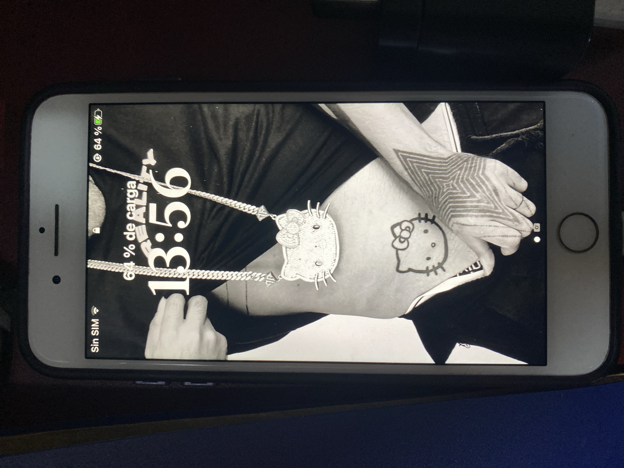 The image shows a cell phone displaying a monochrome image of a person. The person is wearing a necklace with a Hello Kitty pendant. They also have a Hello Kitty tattoo and a geometric star tattoo on their arm. The phone display indicates a time of 3:56 and a battery level of 64%. The phone is an iPhone with a white front and is in a black case. It is not clear if the phone is charging. The phone display language is Spanish. The SIM is not available. The location of the image is not apparent.