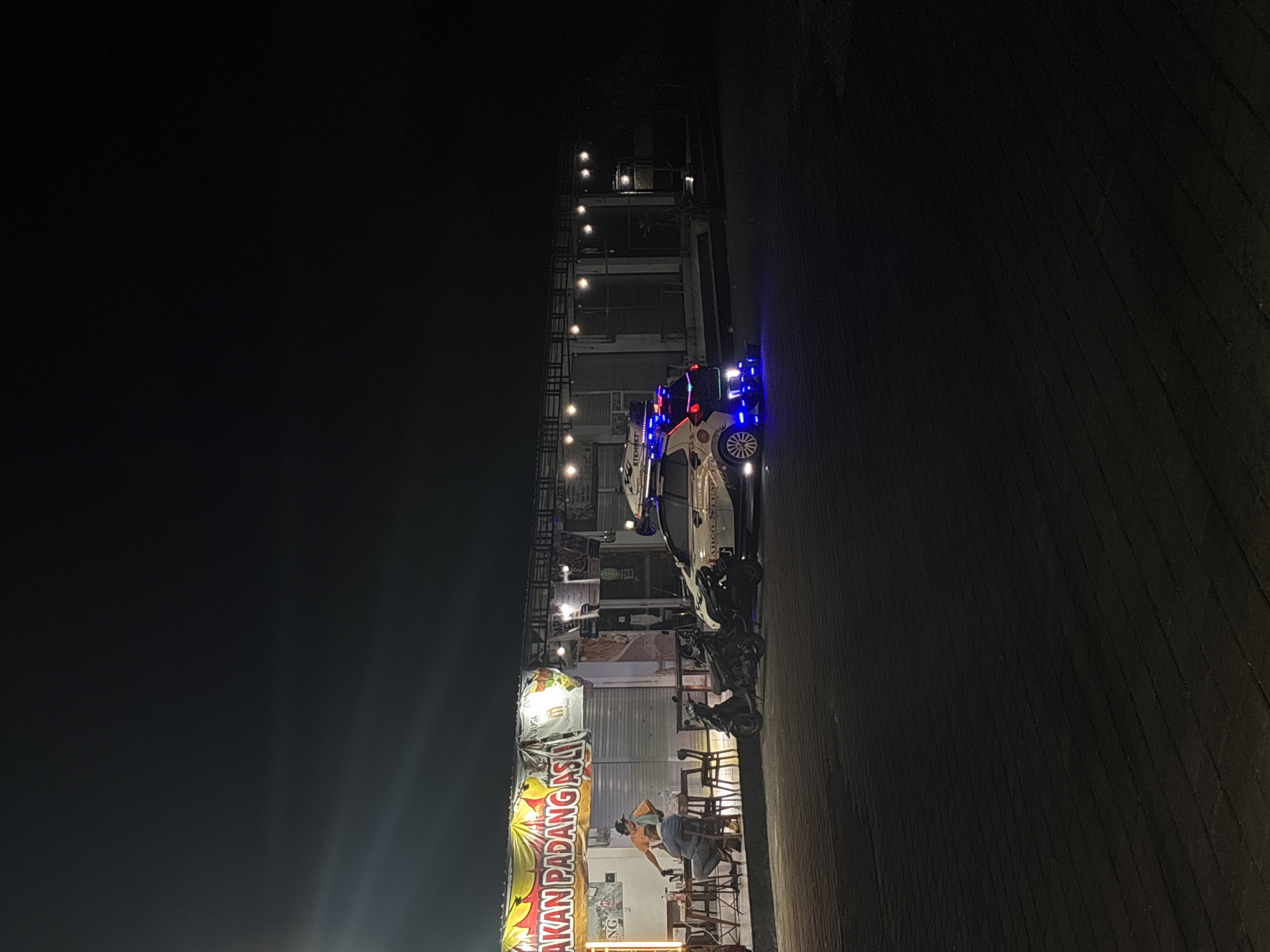 At night, a food stall named "MAKAN PADANG ASLI" is illuminated by bright yellow and white lights, indicating its presence in Kuta Selatan, Indonesia. The stall has a banner with Indonesian text. In front of the stall, two men are seated at tables, one appearing to be an employee serving the other. Several motorcycles are parked to the right of the stall, and a white car modified with blue neon lights is parked in front of a row of shops further down the street. The ground in front of the establishments is paved with dark, interlocking bricks. The overall atmosphere is that of a lively, nocturnal commercial area.