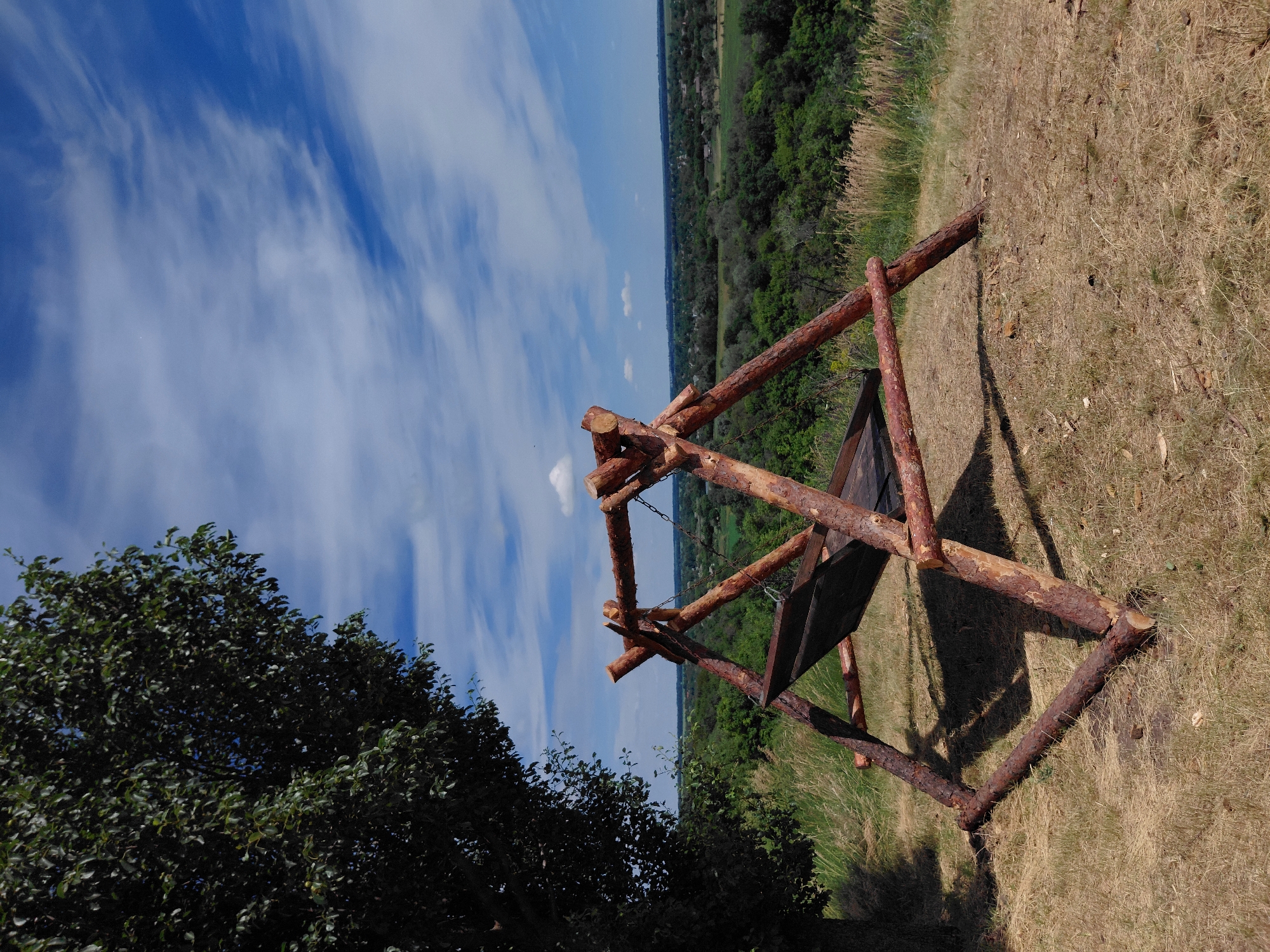 A handmade wooden swing set with a black fabric seat sits on a grassy hilltop overlooking a village in Ustyvytsia, Ukraine. The swing set is constructed from rough-hewn logs, with a triangular frame supporting the seat. A large, dark green tree is to the left of the swing set, its branches reaching towards the sky. The background is a panoramic view of rolling green hills, dotted with houses and trees, under a bright blue sky with wispy white clouds. The ground around the swing set is covered in dry, yellowish grass. The sun casts shadows from the swing set onto the ground.