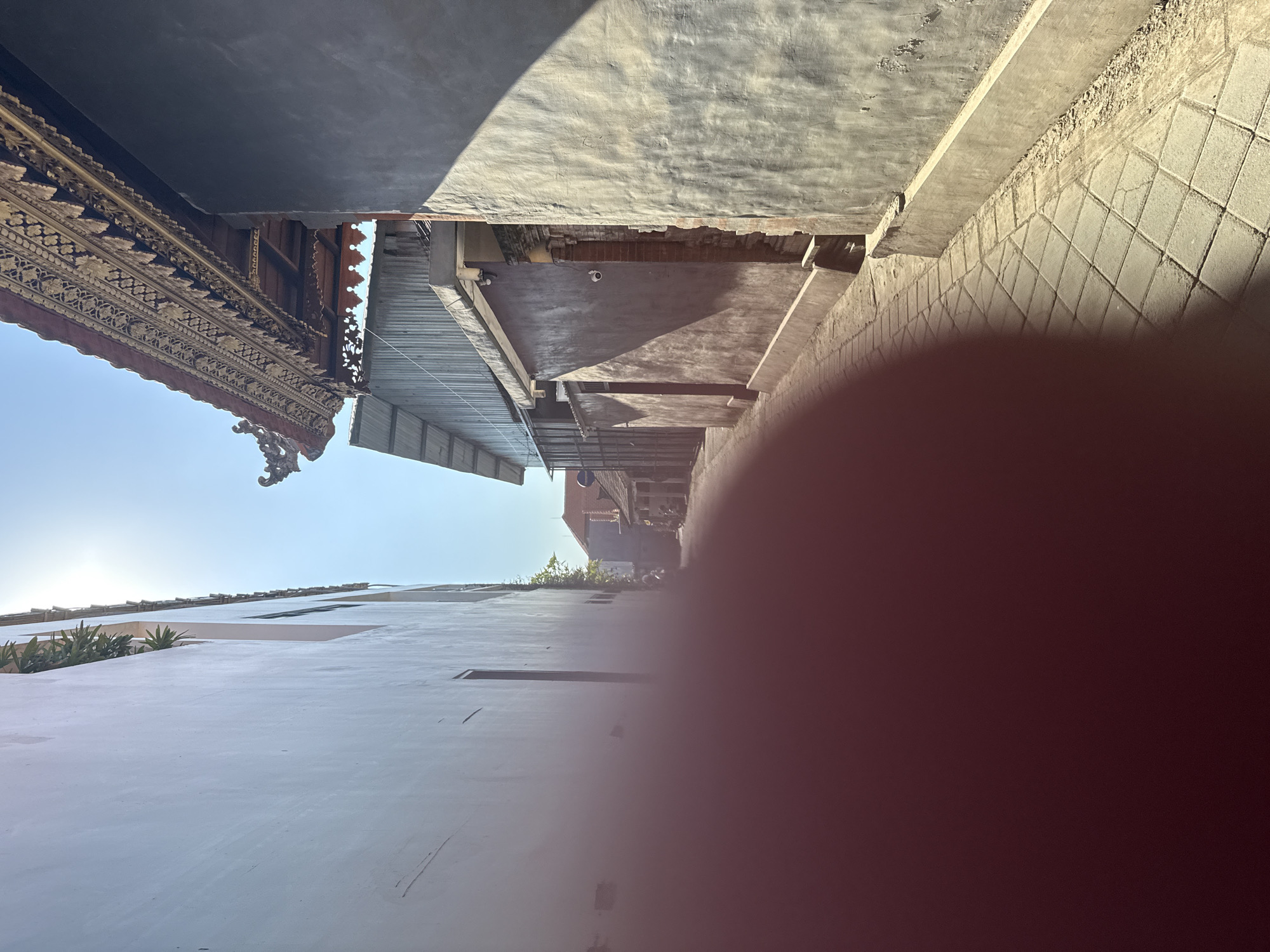 This media file, an outdoor photograph from North Kuta, Indonesia, is presented in a vertical orientation, rotated 90 degrees clockwise. A significant portion of the right side of the image is obscured by a large, blurry, reddish-brown obstruction, likely a finger or thumb covering the camera lens.
Despite the rotation, the scene depicts a narrow alleyway or passage between buildings under bright, clear daylight. On the left side of the depicted pathway (which is the bottom of the image as provided), a tall, light-colored wall rises, featuring some green foliage on a lower ledge. The right side of the pathway (the top of the image as provided) showcases more detailed structures. A prominent roofline with intricate, dark wooden carvings in a traditional, possibly Balinese, style is visible, along with a corrugated metal roof below it. Further back along this side of the alley, concrete walls, some with visible brickwork, are present, and a small surveillance camera is mounted. The ground of the pathway appears to be paved with square-patterned tiles, seen towards the upper right of the image as provided.
The bright sun casts strong, defined shadows across the surfaces, indicating a sunny day. The sky, visible between the buildings, is a clear blue. No people or specific activities are visible, presenting a static street scene. There is no discernible text in the photograph.