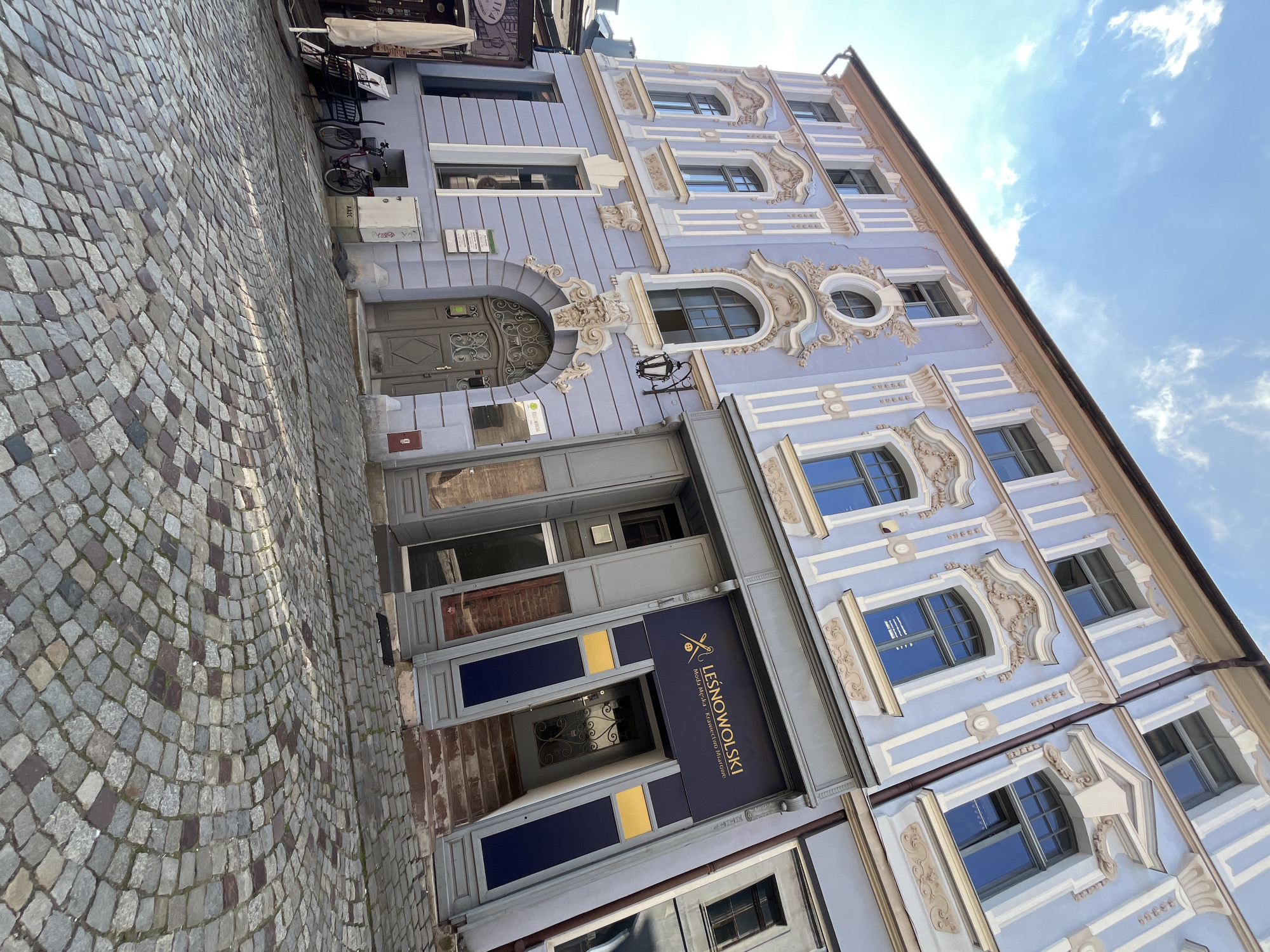 The image shows a close-up of a light purple building with ornate decorative elements around its windows. The building has multiple stories and is situated on a cobblestone street. The architecture suggests a historical European setting. The building has several windows, some of which are arched and decorated with stucco. There is a prominent entrance with a sign that reads "LEŚNOWOLSKI" in gold lettering, along with smaller text underneath. Two bicycles are parked near the entrance. The sky above is blue with scattered white clouds, indicating it is daytime and likely fair weather. The angle of the photograph is tilted, giving a dynamic perspective of the building and street. The scene appears to be a quiet street in a city or town.