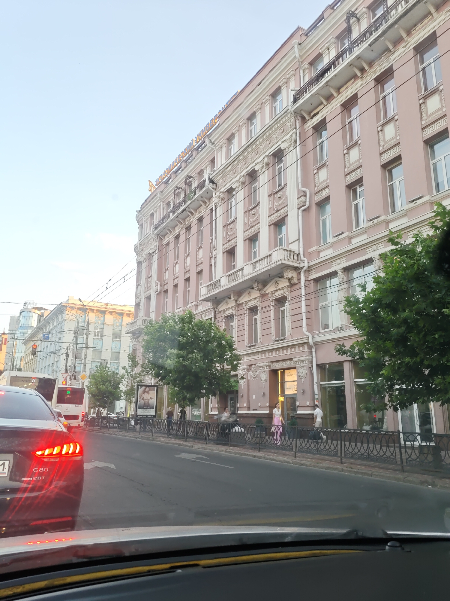 The image captures a view from inside a vehicle looking out at a street scene in Rostov-on-Don, Russia. The dominant feature is a large, ornate, pink building with multiple stories and detailed architectural elements, including balconies, columns, and decorative moldings. Golden lettering is visible on the top of the building.
In the foreground, the rear of a dark-colored car is visible, its red taillights illuminated. The car has a license plate that begins with "G80". Further down the street, several buses are stopped. One bus has the number "39" displayed. People are visible walking on the sidewalk alongside the pink building, some standing near an entrance and a railing.
The street is paved, and overhead wires suggest it's a route for public transport like trams or trolleybuses. Trees with green foliage line the sidewalk. A traffic light is visible in the distance. The sky is overcast. The overall impression is of a typical urban street scene on a daytime, likely overcast day.