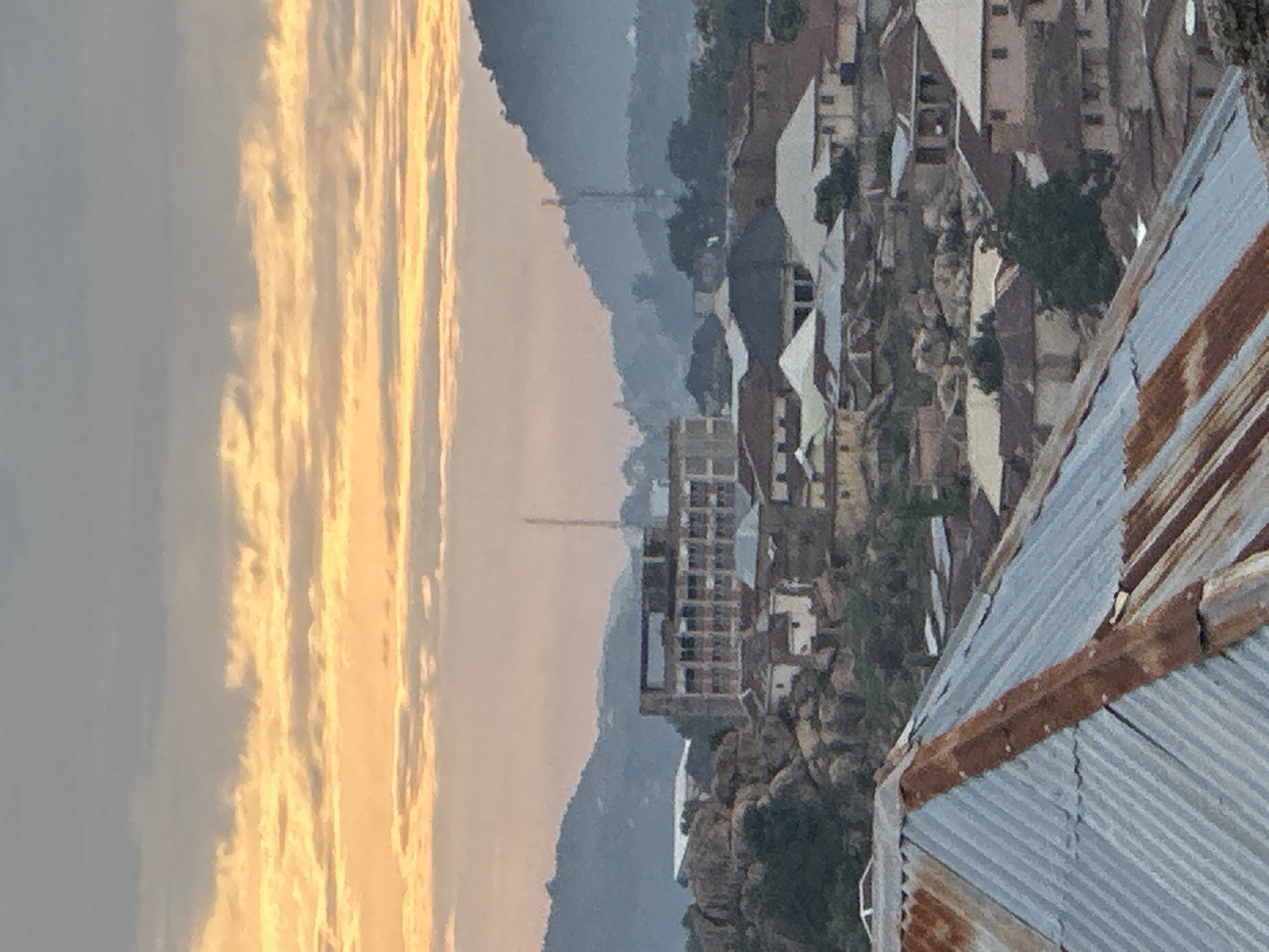 This image presents an elevated panoramic view of a densely populated urban settlement in Jos, Nigeria, under crepuscular light conditions, likely dawn or dusk. The sky, occupying the upper third of the frame, features distinct horizontal bands of orange-yellow clouds indicative of either sunrise or sunset, contrasted with a softer grey-blue above and below.
In the immediate foreground, the steeply sloped section of a corrugated metal roof is visible, characterized by prominent rust and weathering. The roof surface is a mix of dull silver-grey and significant patches of reddish-brown rust, particularly along the ridges and seams.
The midground is dominated by a compact urban landscape. Numerous residential buildings, primarily single or two-story, are densely packed together, many featuring similar corrugated metal roofs to the foreground, showing varying degrees of rust and wear. Buildings are constructed with light-colored walls, possibly painted or rendered. The built environment is integrated with a rugged natural terrain, featuring numerous large, exposed rock formations and boulders scattered amongst the structures. A prominent multi-story concrete building, appearing either partially constructed or in a state of disrepair with visible structural frames and window openings, stands centrally within this urban cluster. Sparse green vegetation, consisting of trees and shrubs, is interspersed throughout the rocky and built-up areas. No individuals are visible within the scene.
The background reveals a series of distant hills and mountains, rendered in muted blue-grey tones due to atmospheric haze, suggesting significant distance. Two tall, slender structures, likely telecommunication masts or antennas, are discernible on separate hilltops towards the left and right sides of the background.