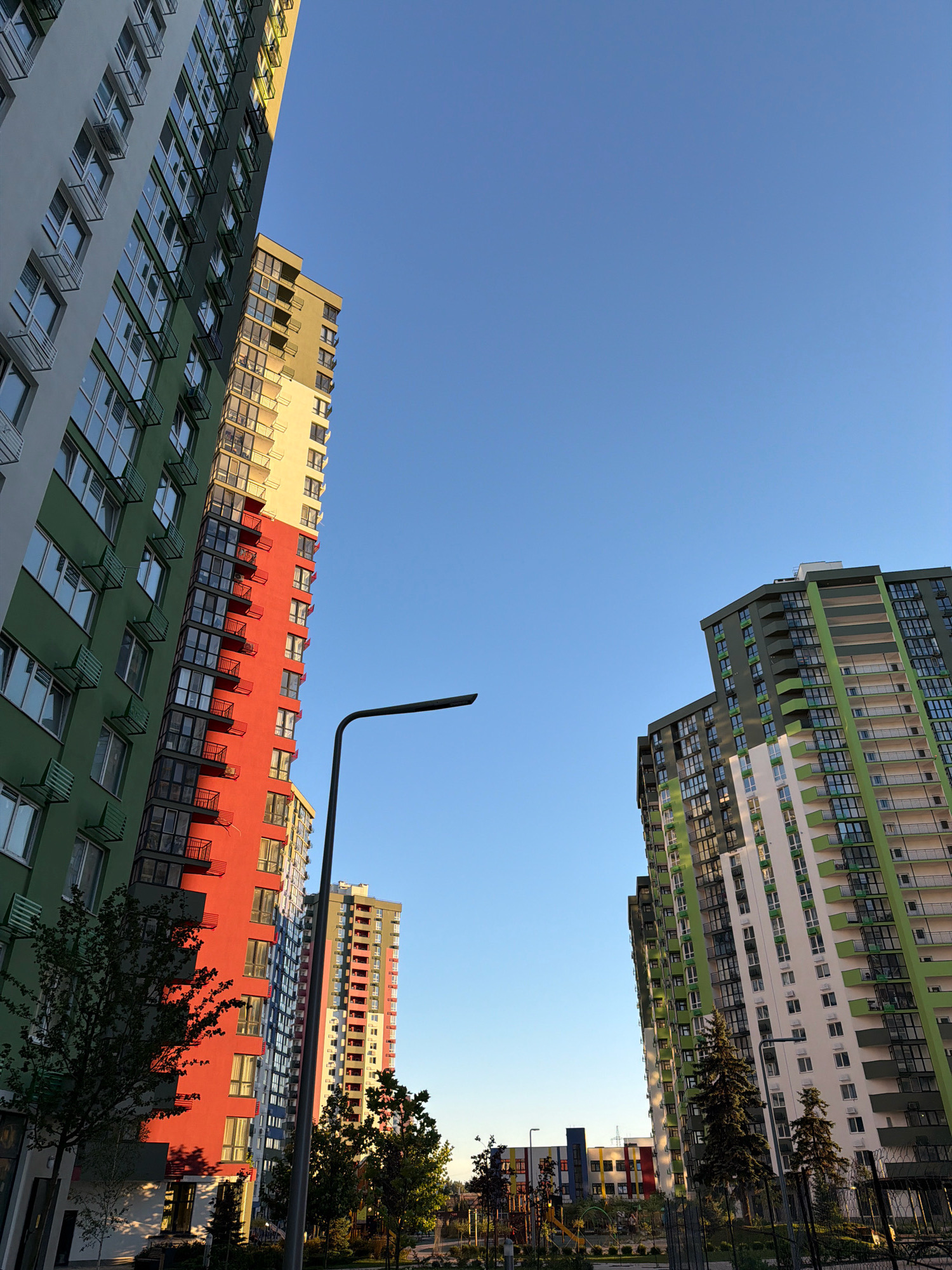 The photograph captures a view of modern residential buildings in Kyiv, Ukraine, under a clear blue sky. The buildings are tall, multi-story structures with a striking color palette of green, red, yellow, and white facades. Numerous windows and balconies are visible, with some balconies featuring railings and exterior air conditioning units.
In the foreground, a dark grey streetlamp with a curved arm extends into the frame from the left. To the left of the streetlamp, a portion of a building with a green and white facade is visible, with small trees and their shadows cast on the lower part of the building.
In the mid-ground, several tall residential buildings are arranged. One building prominently features a section painted bright red, contrasted with sections of yellow and green. Another building further to the right, also a tall residential complex, is primarily decorated with green and white sections, with balconies visible along its length.
In the background, more buildings are visible, including a lower, colorful building with sections of blue, yellow, red, and white, suggesting it might be a different type of structure or part of another complex. A playground area with some equipment can be seen in the far background. Tall, dark green coniferous trees are interspersed between the buildings. The overall impression is of a contemporary urban residential area.