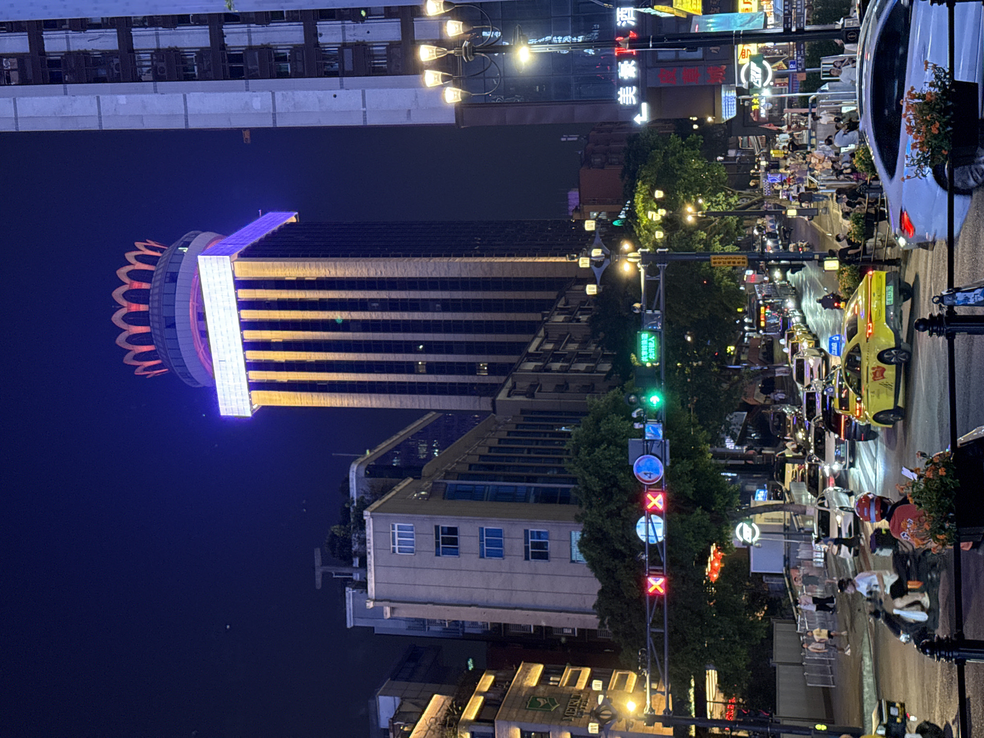 This is a nighttime, eye-level, medium shot of a busy street scene in Yuzhong District, China. Tall buildings with illuminated windows line the street, some featuring prominent signage. A distinctive tower building with a crown-like illuminated top stands out. Traffic lights, including red X's and a blue circle with a white number 4, are visible above the intersection. Numerous vehicles, including cars and motorcycles, are present on the road, some appearing to be in traffic. Pedestrians are walking on sidewalks and crossing the street. Streetlights and the lights from buildings and vehicles illuminate the scene, creating a vibrant, urban nighttime atmosphere. Some of the buildings have green trees and shrubbery in front of them.
