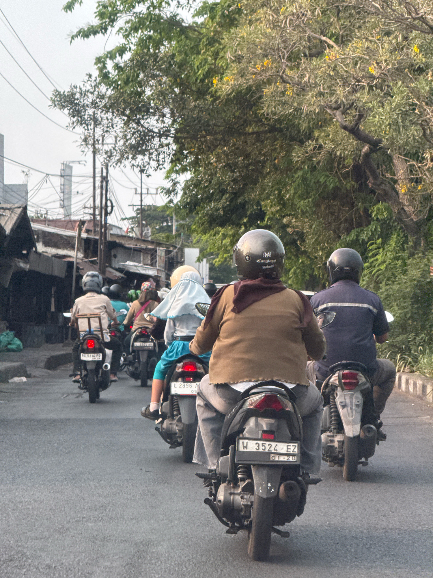 This is a medium-shot, eye-level, outdoor, daytime photograph taken from behind, capturing several people riding motorcycles on a street in Surabaya, Indonesia. In the foreground, a person wearing a brown jacket and a dark helmet is riding a motorcycle with the license plate "W 3524 EZ". To their right and slightly ahead, another person in a dark blue shirt and dark helmet is riding a motorcycle, with their license plate partially visible. Further back and to the left, a person in a light brown jacket and a silver helmet is on a motorcycle with the license plate "BG 5214 FO". There are at least two other motorcycles visible in the mid-ground, one with a passenger wearing a light blue head covering and a white and blue outfit. The street is paved and appears to be an urban environment with buildings and market stalls lining the left side of the road. Lush green trees with some yellow flowers are visible on the right side of the frame, and power lines are strung across the sky. The overall scene depicts traffic on a street in Surabaya.