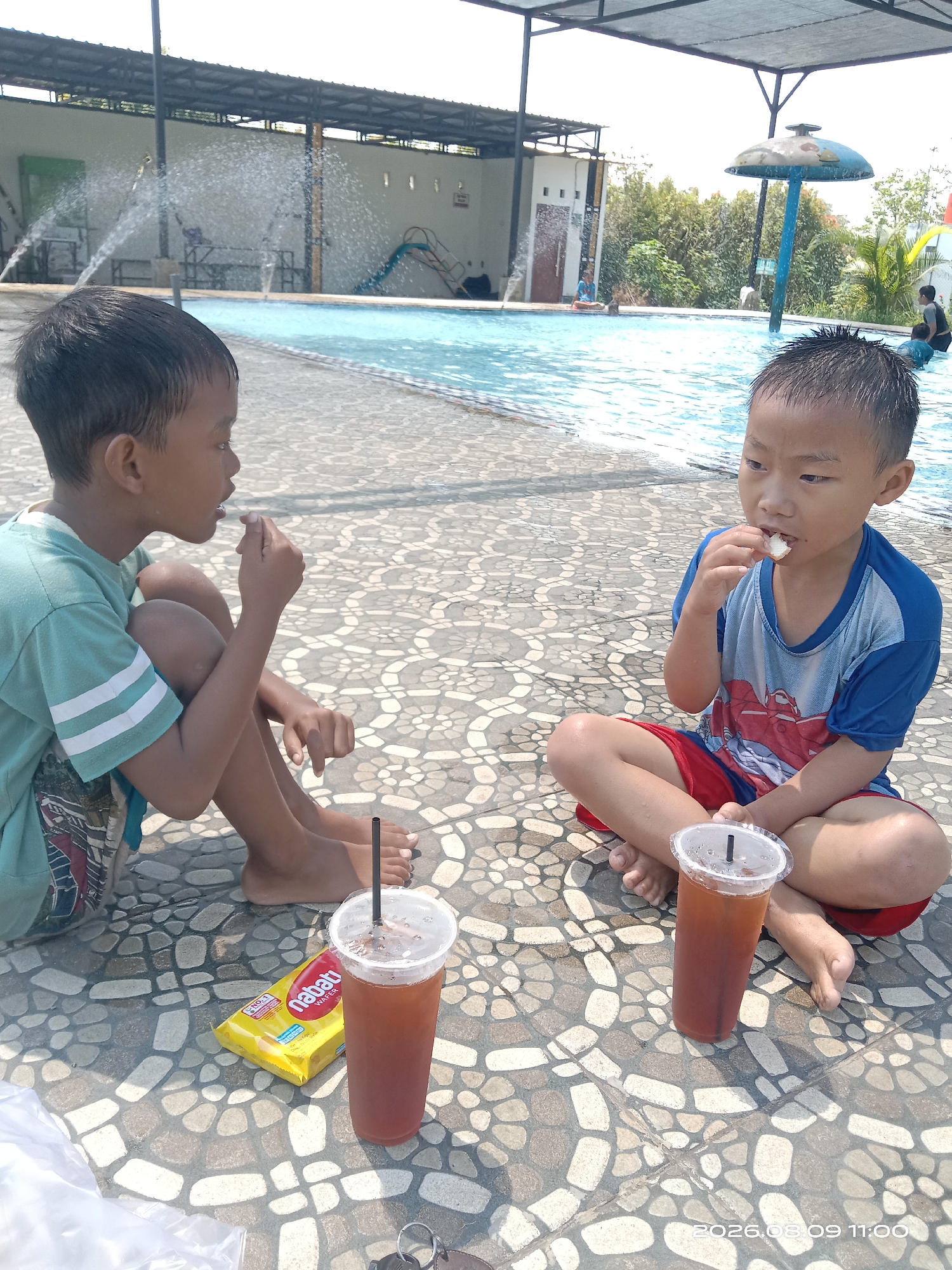 Two young boys are sitting on the tiled ground next to a swimming pool in Blimbing, Indonesia. The boy on the left is wearing a light green t-shirt with white stripes on the sleeves and dark shorts. He is facing towards the boy on the right and appears to be eating. The boy on the right is wearing a blue t-shirt with a red graphic and red shorts. He is also facing the other boy and is eating something. In front of them are two plastic cups with dark liquid and straws, and a yellow package of "nabati Wafer". The swimming pool is visible in the background, with water fountains spraying. The ground is paved with decorative tiles. The image is taken during the day with bright sunlight. The timestamp at the bottom right corner indicates "2026.08.09 11:00".