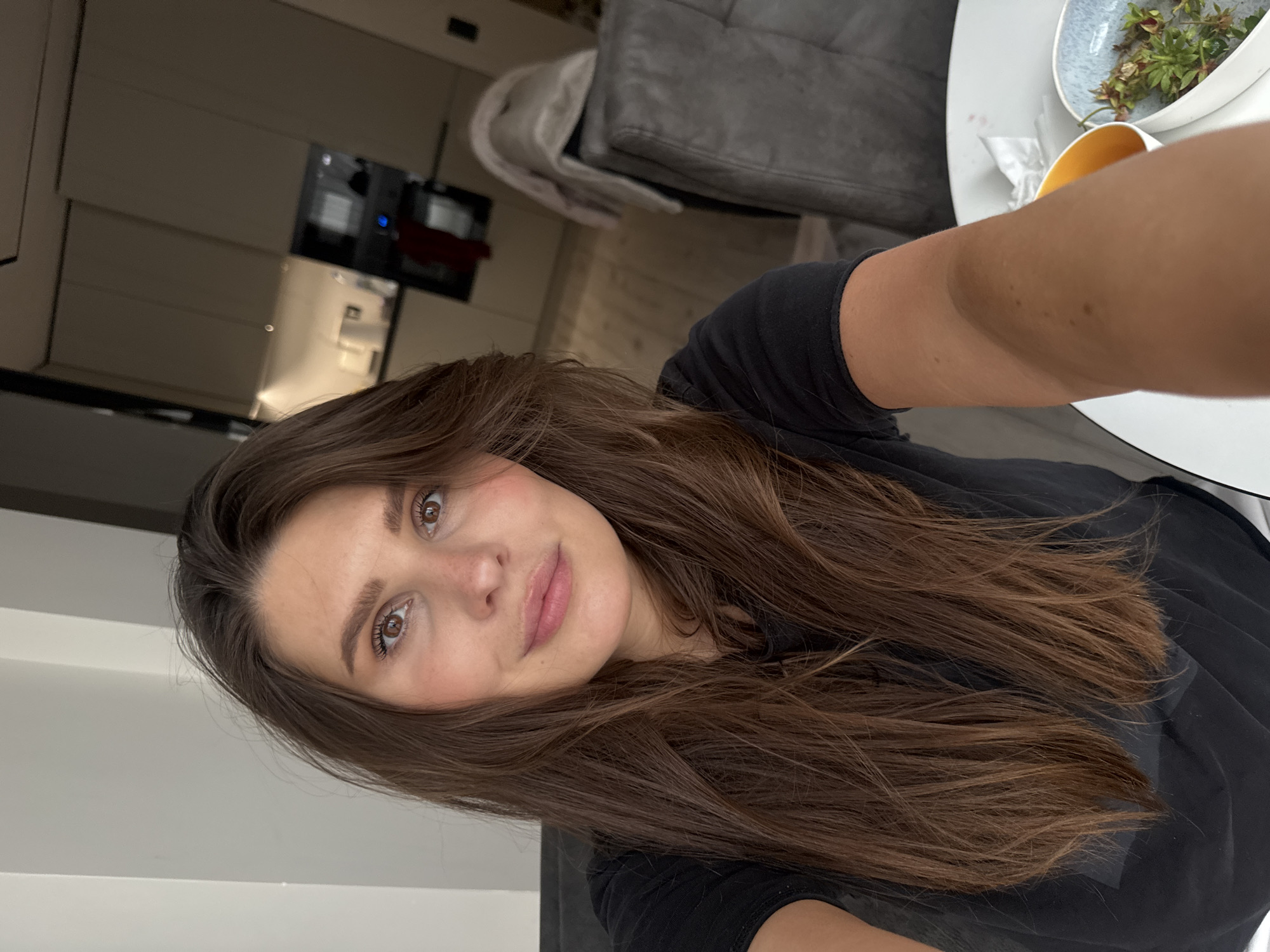 A lone female adult with long, brown hair and brown eyes is depicted in a close-up selfie, captured indoors within what appears to be an apartment or house located in Sofiivska Borshchahivka, Ukraine. She wears a black t-shirt and displays a neutral to slight smile. Her right arm is visible in the lower right foreground. In the immediate foreground, a white circular table holds a yellow cup with an orange liquid, a blue-patterned bowl containing green plant material, and a white tissue. The background reveals an open-plan living area and kitchen, featuring a dark grey upholstered chair with a light-colored throw, light wooden flooring, and modern kitchen cabinetry. The kitchen area includes white flat-front units, a white kettle, and an oven or microwave unit with a red object partially visible inside. The space is illuminated by overhead and under-cabinet lighting, with a white wall section visible on the left.