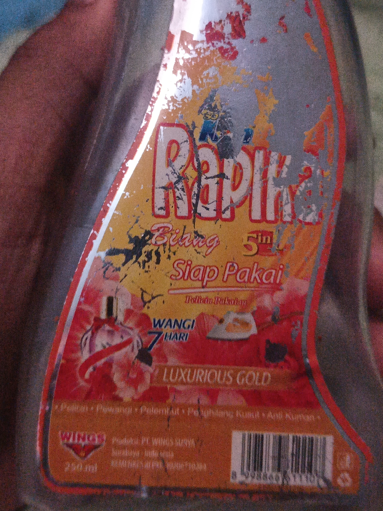 The image shows a close-up of a damaged label on a bottle, likely a fabric softener, with Indonesian text and a barcode. The label features the brand name "Rapika" in large, red and white letters. Below that, smaller text reads "Bihing" and "Siap Pakai" which translates to "Ready to Use." There is also text indicating "Wangi 7 Hari" meaning "Fragrant for 7 Days." A section at the bottom displays "LUXURIOUS GOLD" and a list of benefits including "Pelembut" (Softener) and "Anti Kuman" (Anti-Germs). The brand logo "WINGS" is visible in red and white. The barcode and product size "250 ml" are also present. The label is peeling and worn, with scuffs and discoloration indicating age and use. The background is out of focus, showing a blurred hand or arm holding the bottle. The image does not provide information about a specific location in Temuwulan, Indonesia, nor does it depict any specific scene within the city.