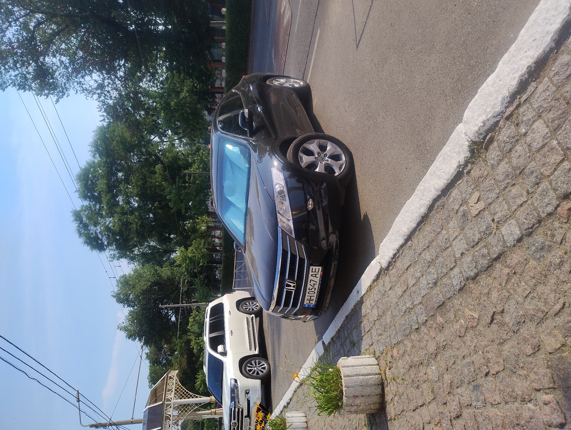 Two cars are parked on a street with trees and a blue sky in the background. The car in the foreground is a black Honda sedan, license plate HH 0547 AE. To its left is a white Toyota SUV, license plate HH 9522 PC. The street is paved with cobblestones and asphalt. There are potted plants on the left side of the street.