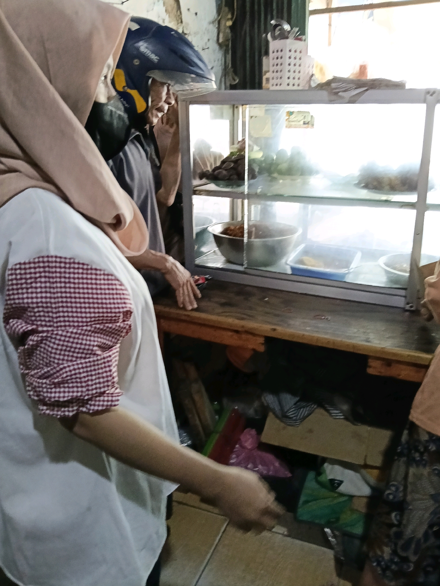In the city of Belimbing, Indonesia, a scene unfolds at what appears to be a food stall. The focal point is a glass display cabinet containing various food items. Two individuals are partially visible. One is wearing a light-colored hijab and a white shirt with red and white checkered sleeves. Their arm is extended, and their hand is near the edge of the cabinet, possibly interacting with something. The second individual, wearing a blue helmet with yellow accents and a dark shirt, is also near the cabinet, looking towards it. The environment surrounding the stall is somewhat cluttered, with shelves filled with items, bags, and what looks like building materials in the background. The lighting suggests it is daytime, with natural light filtering in from an opening.