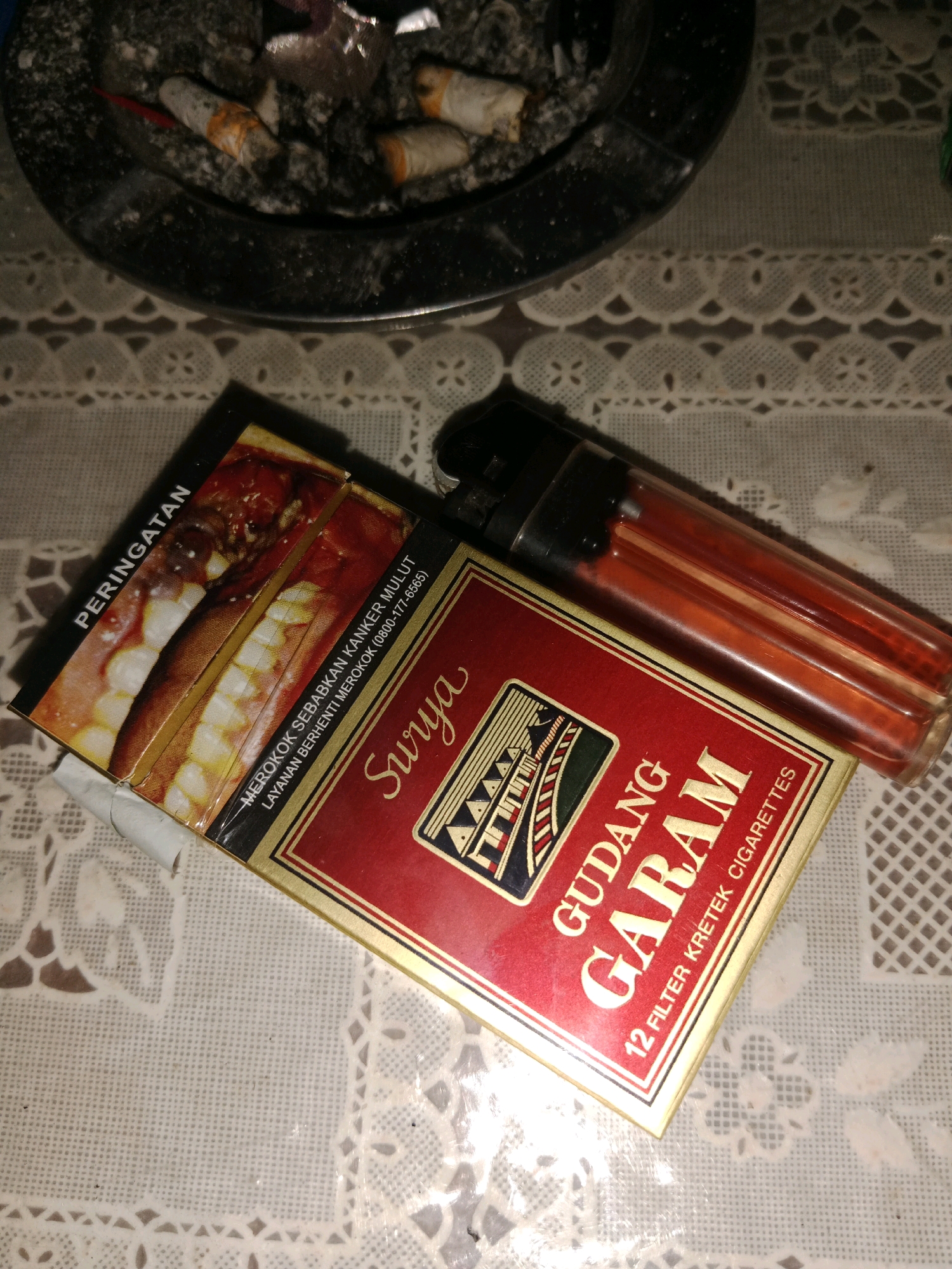 The image shows a close-up of a table covered with a white, patterned lace tablecloth. On the table are a pack of Gudang Garam Surya cigarettes, a transparent lighter with red fluid, and an ashtray containing several cigarette butts. The ashtray is in the upper portion of the image, and the cigarette pack and lighter are in the lower right. The pack of cigarettes is red and gold with text and images. The lighter is resting on top of the cigarette pack. The scene appears to be indoors, possibly in a home or a casual setting in Ringinrejo, Indonesia.