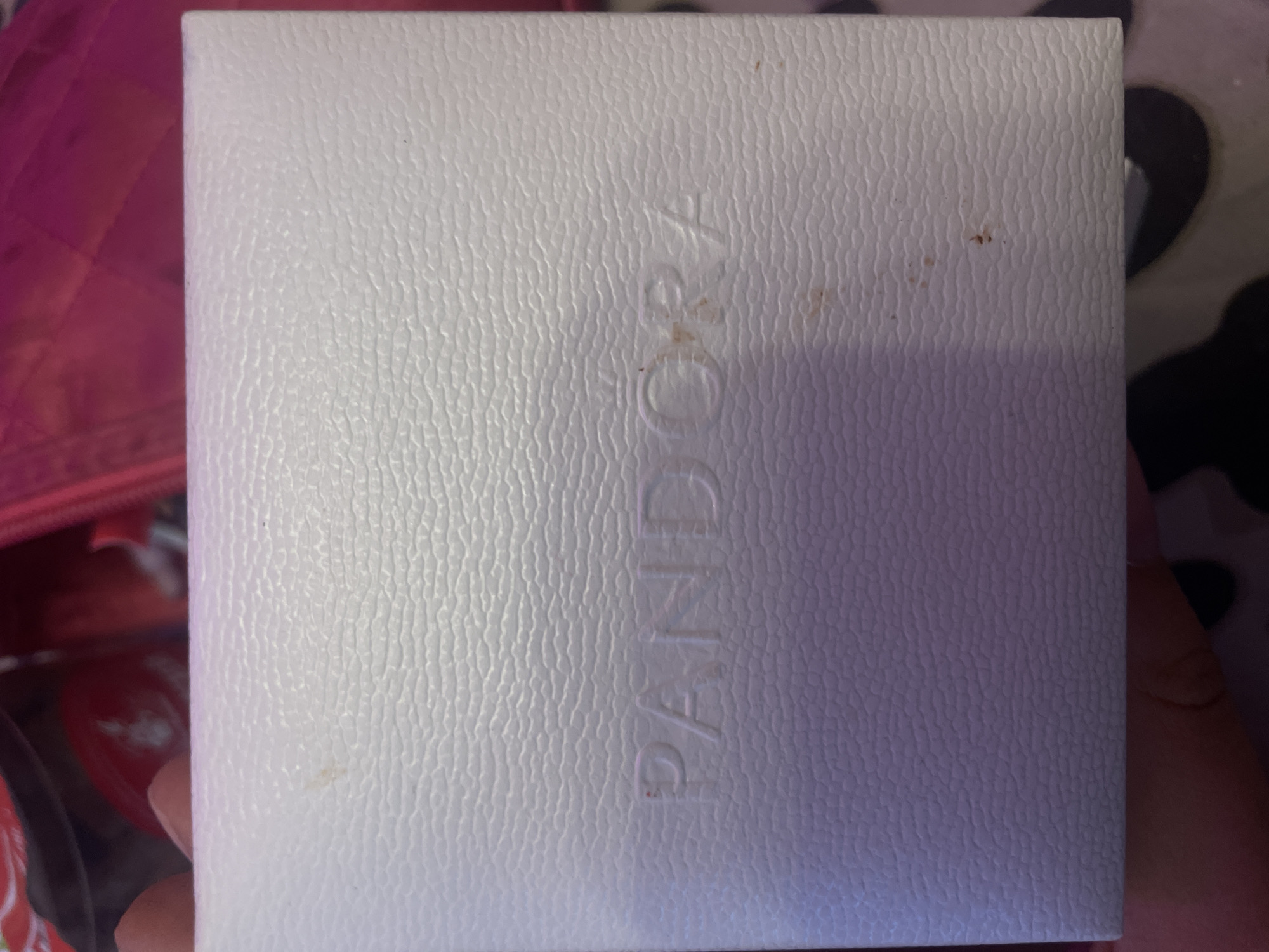 The image shows a white Pandora jewelry box. The brand name "PANDORA" is embossed on the lid. There are a few small brown smudges on the lid. The box appears to be made of textured material. The background is out of focus, but appears to be a pink quilted fabric. The image is taken from a top-down angle, with a hand visible at the bottom of the frame. The scene is indoors. There is no information to suggest this is in Ladozhskoe Ozero, Russia.