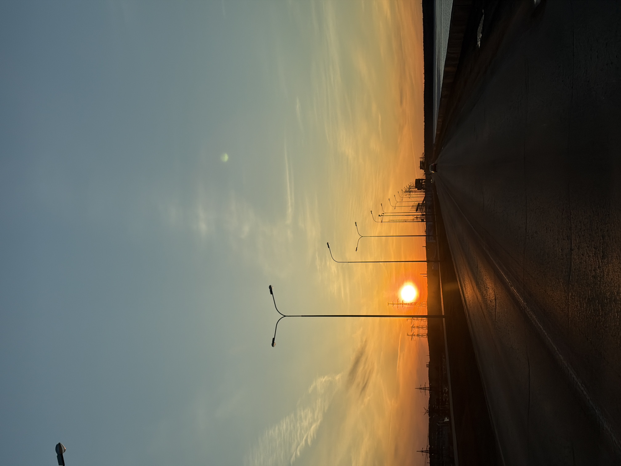 The image depicts a sunset over a road in Chaikovsky, Russia. The sun is positioned on the horizon, casting a warm glow on the scene. Streetlights line the road. Power lines are visible in the background. The sky transitions from a clear blue to an orange hue near the horizon. The road surface is dark, reflecting the sunlight. The image is rotated.