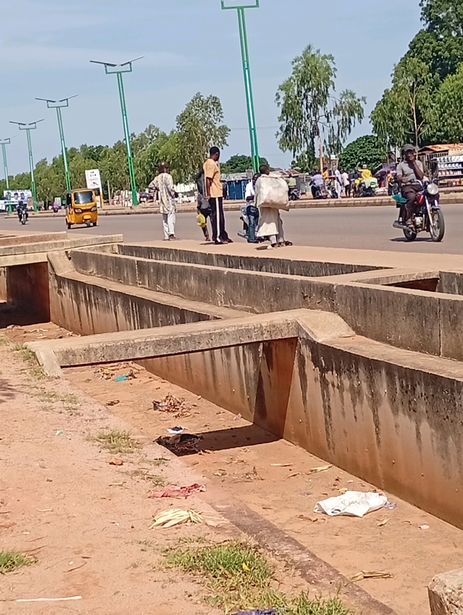 The image shows a street scene in Tumfure, Nigeria. The foreground is dominated by a concrete drainage ditch, which is dry and contains some litter, including a white plastic bag. Behind the ditch is a paved road with several people walking and riding motorcycles.
On the road, a yellow auto-rickshaw is visible, as well as several motorcycles. A man is riding a motorcycle in the right side of the image, wearing a grey shirt and a cap. Three men are walking on the sidewalk in the middle of the image. One man in a white and black patterned outfit is carrying a white sack over his shoulder. Another man in a yellow shirt is walking behind him. A third man in a white garment is walking towards the left. Further down the road, more people and vehicles are visible.
In the background, there are trees and buildings. Several streetlights with solar panels are erected along the road. The sky is clear and blue, indicating a sunny day. The overall scene depicts a typical urban street in Nigeria, with a mix of pedestrian and vehicular traffic, along with infrastructure like drainage systems and streetlights.